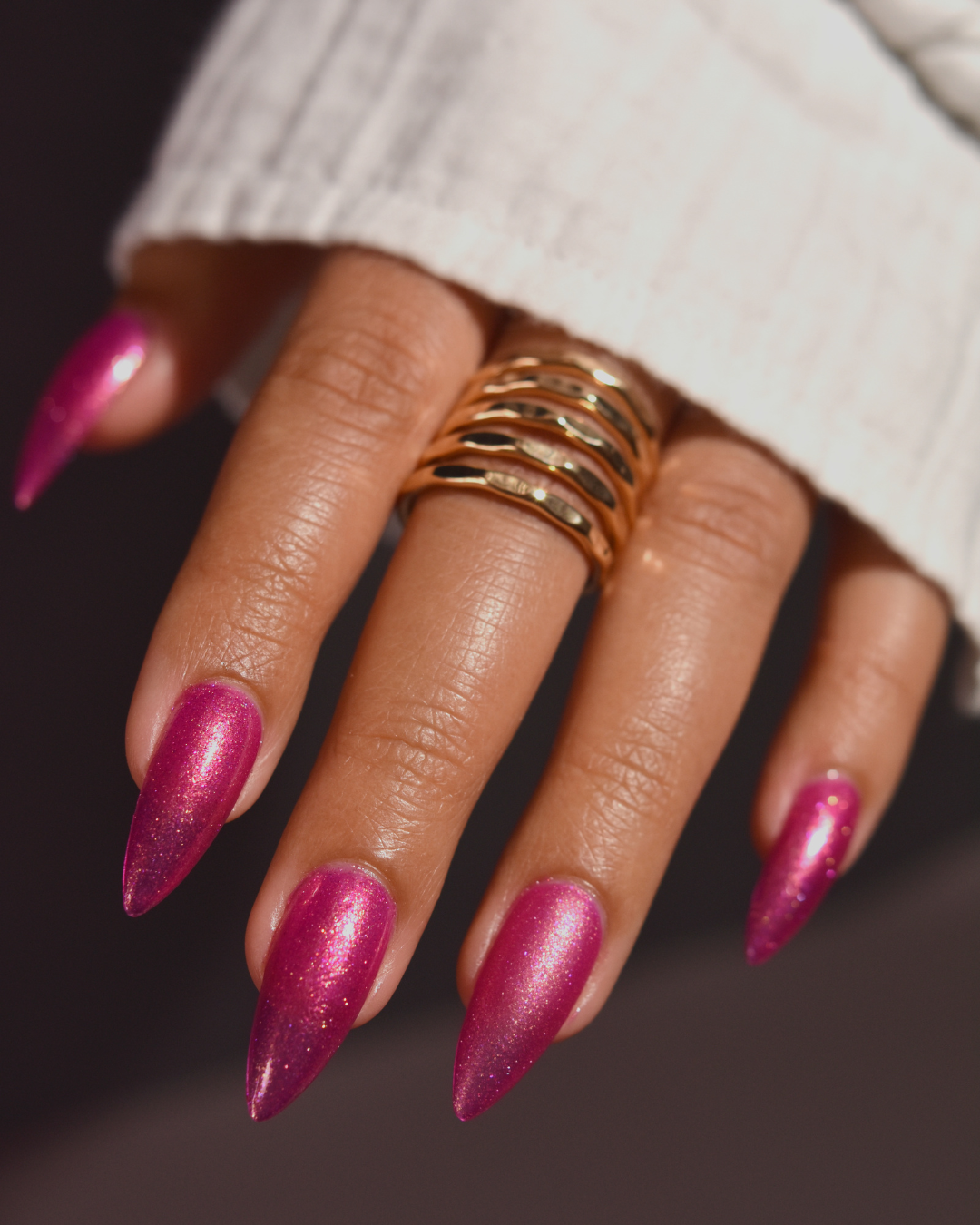 A close-up of a hand with long, glossy magenta stiletto nails and a stack of gold rings, wearing a white ribbed knit sleeve against a dark background.