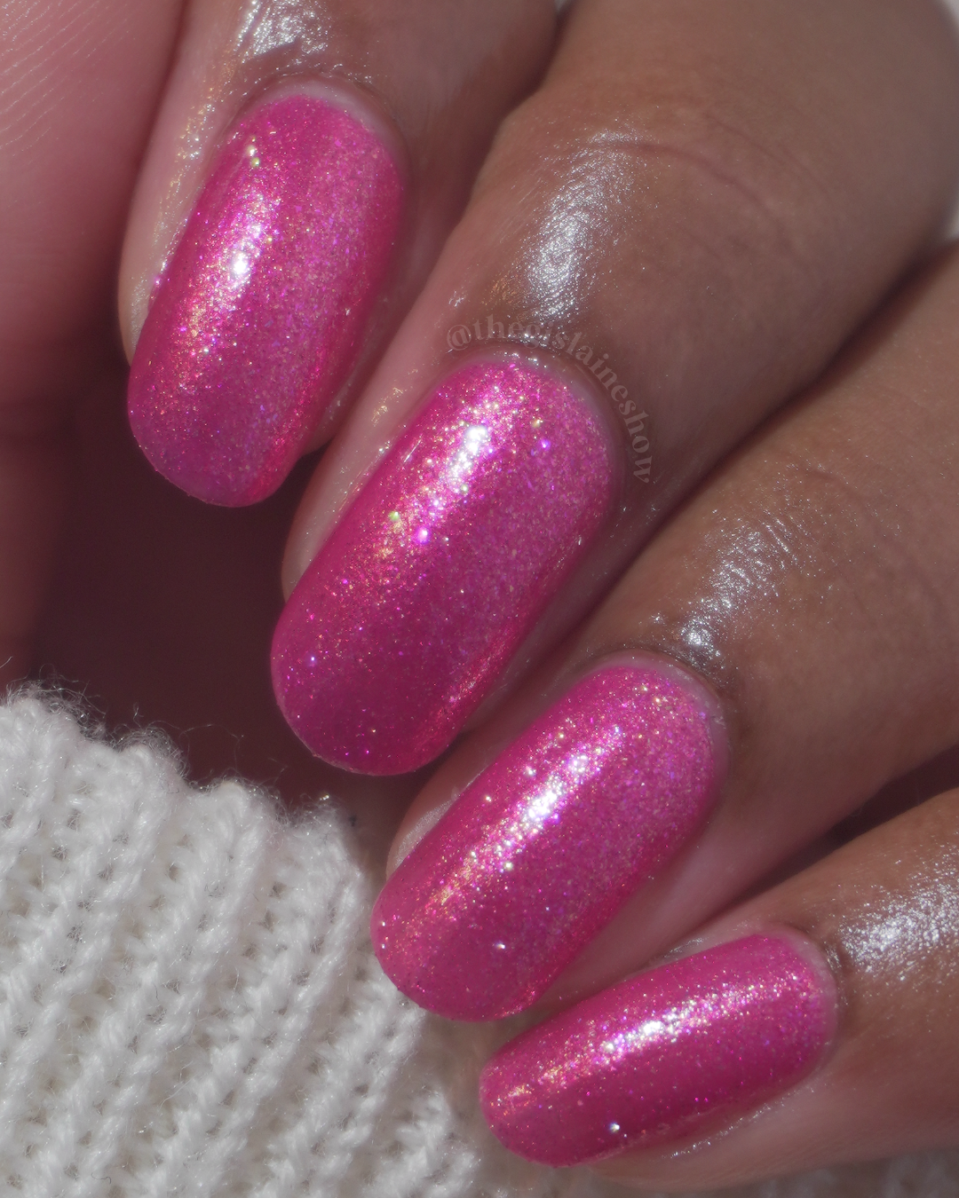 A close-up of a hand with fingernails painted in a bright pink glitter polish, resting against a white knit fabric.