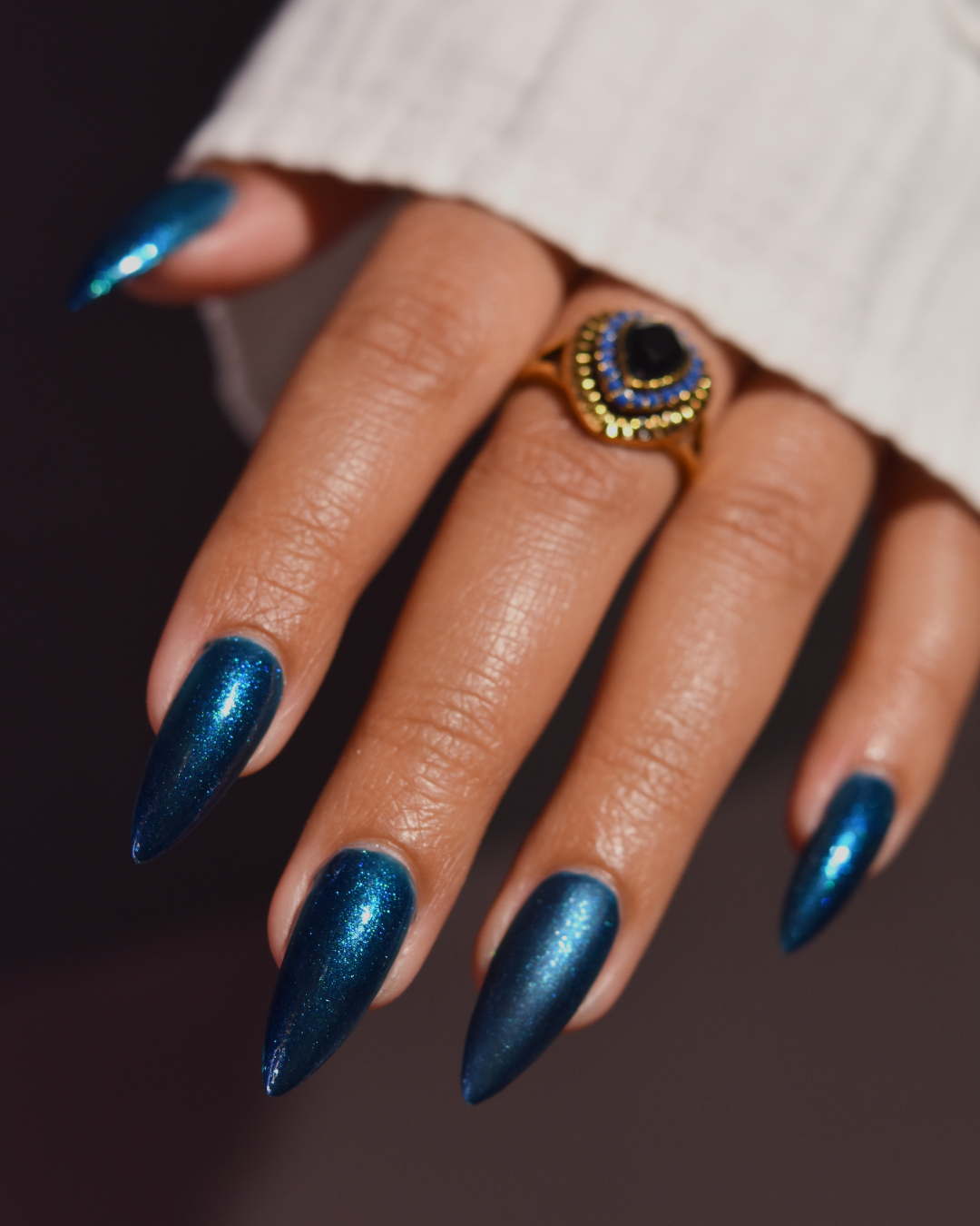 A close-up of a hand with long, stiletto-shaped nails painted in a metallic deep blue shade, wearing a gold ring with a black and blue stone. The hand is resting against a white ribbed sleeve, set against a dark, solid background.