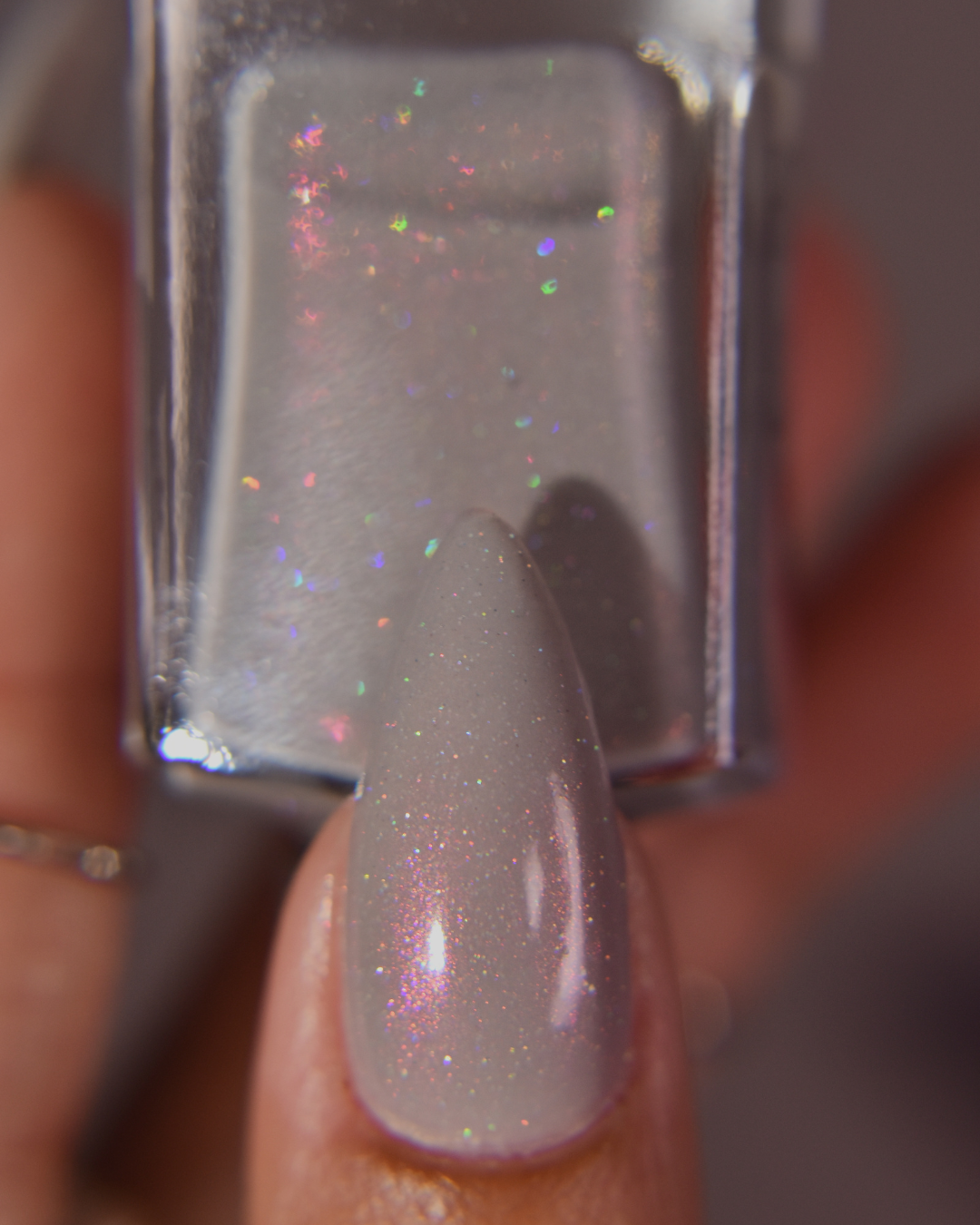 A close-up of a fingernail painted with a sheer light gray nail polish containing fine iridescent holographic glitter, held in front of a matching nail polish bottle.