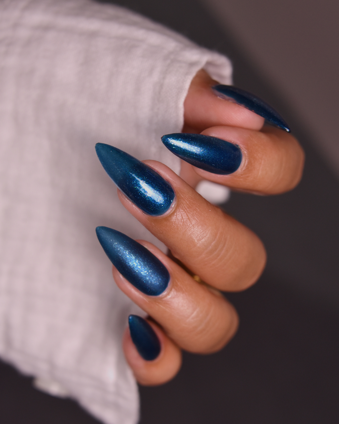 A close-up of a hand with long, stiletto-shaped nails painted in a metallic deep blue shade, resting against a light-colored textured sleeve.