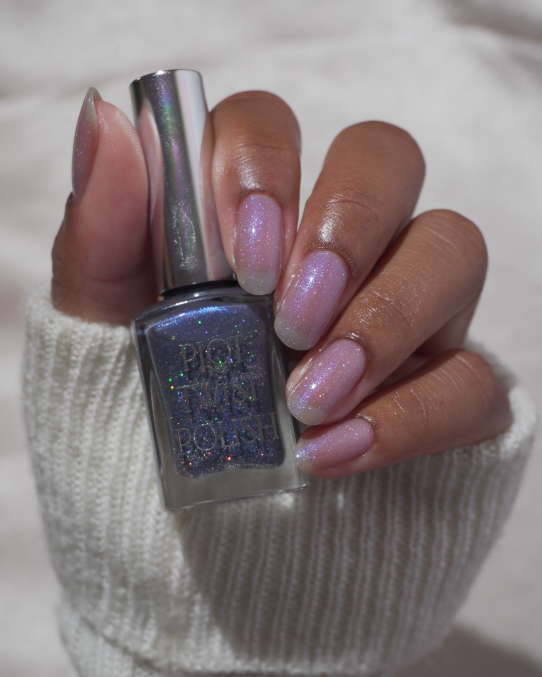 A manicured hand with a translucent shimmer nail polish holding a blue nail polish bottle.