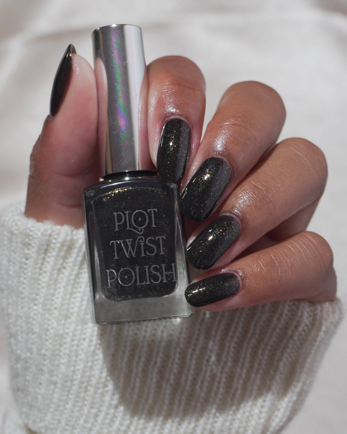 A manicured hand with a black polish with gold flakes holding a bottle of polish.