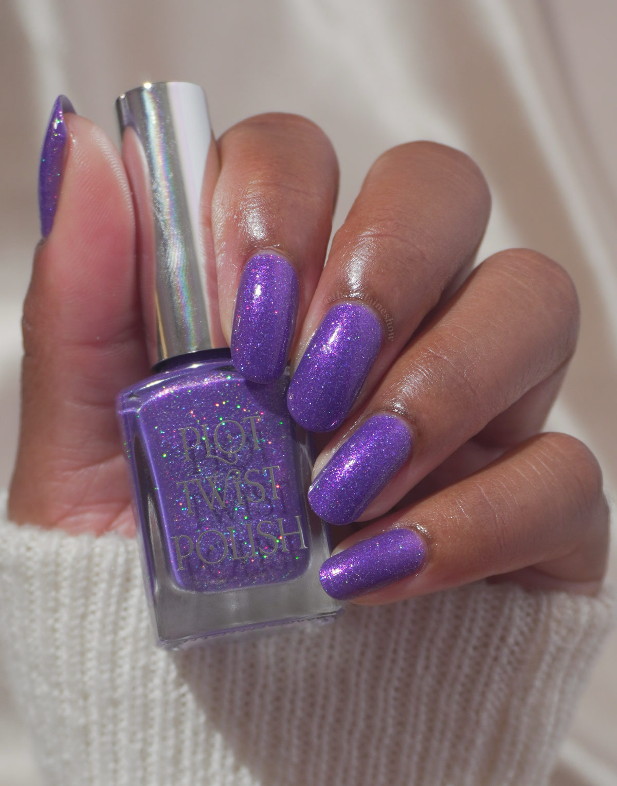 A hand with long, glossy purple glitter nails holds a bottle of matching purple nail polish against a soft cream-colored background.
