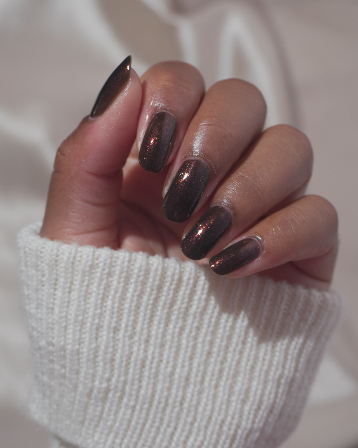 A close-up of a hand with manicured nails painted in a dark metallic bronze-brown polish, wearing a white ribbed knit sweater against a soft light background.