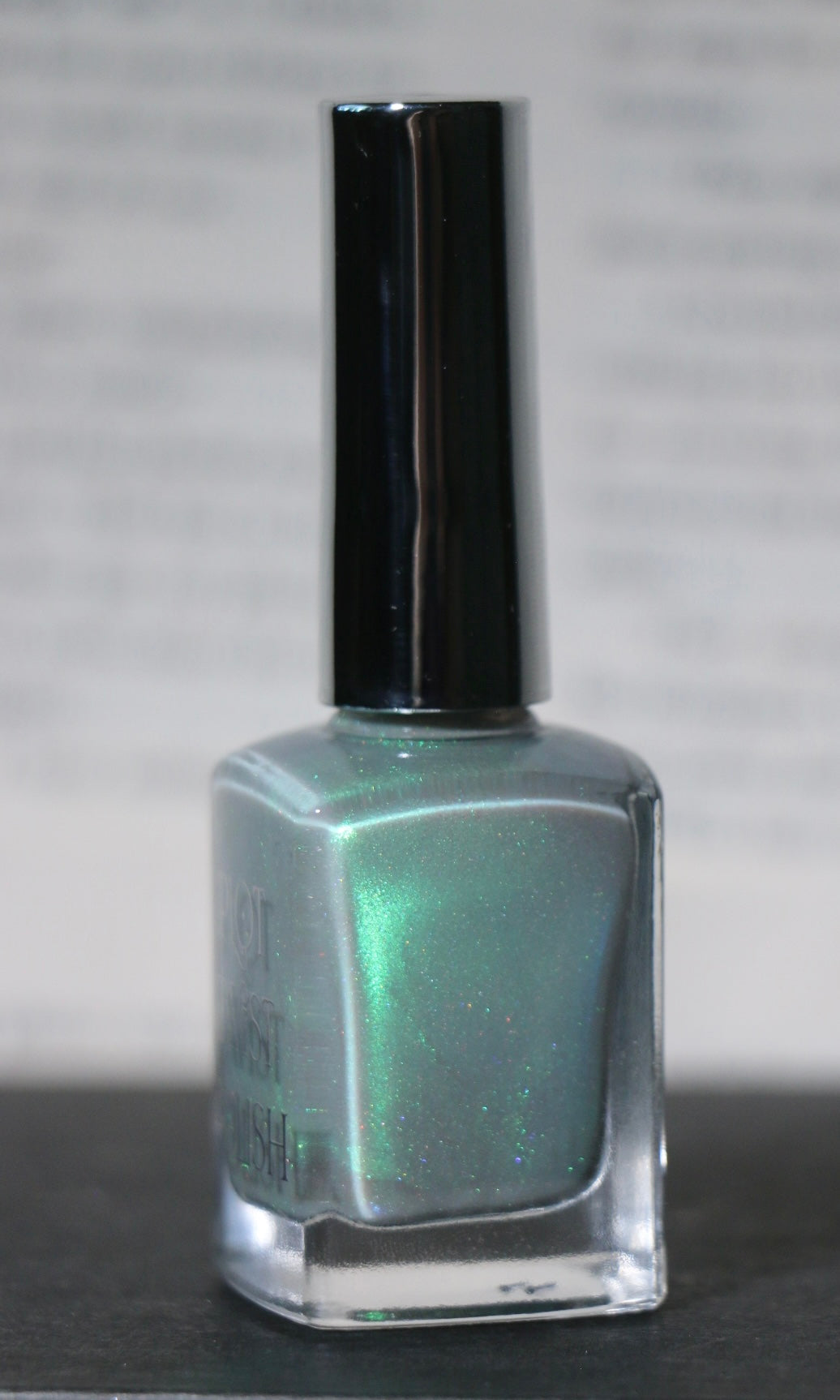 A bottle of Plot Twist Polish nail lacquer in a shimmering grey green green shade, featuring a silver cap and clear glass bottle.