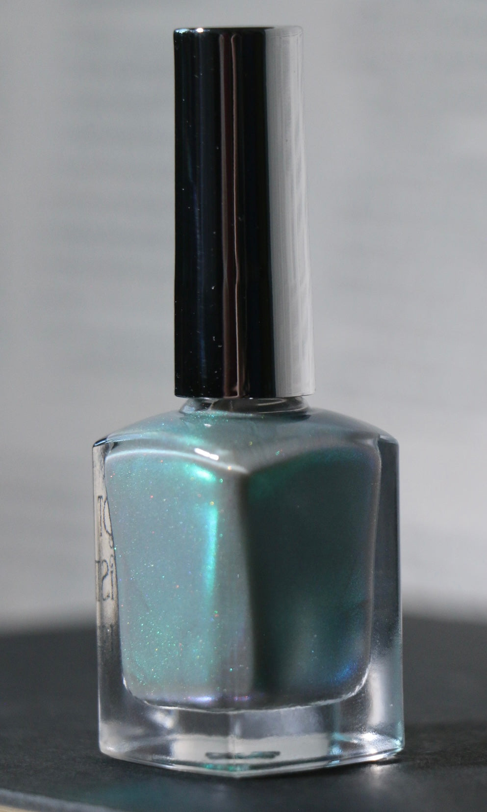 A bottle of Plot Twist Polish nail lacquer in a shimmering grey green green shade, featuring a silver cap and clear glass bottle.