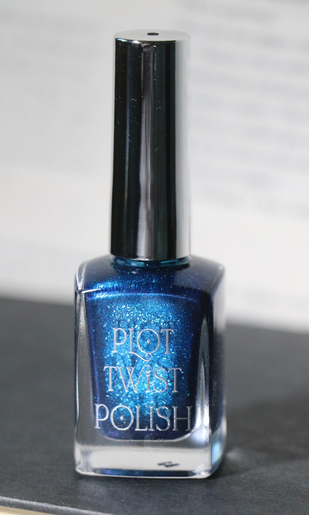 A bottle of blue glitter nail polish with a silver cap, labeled 'PLOT TWIST POLISH', resting on a dark surface against a light gray background.
