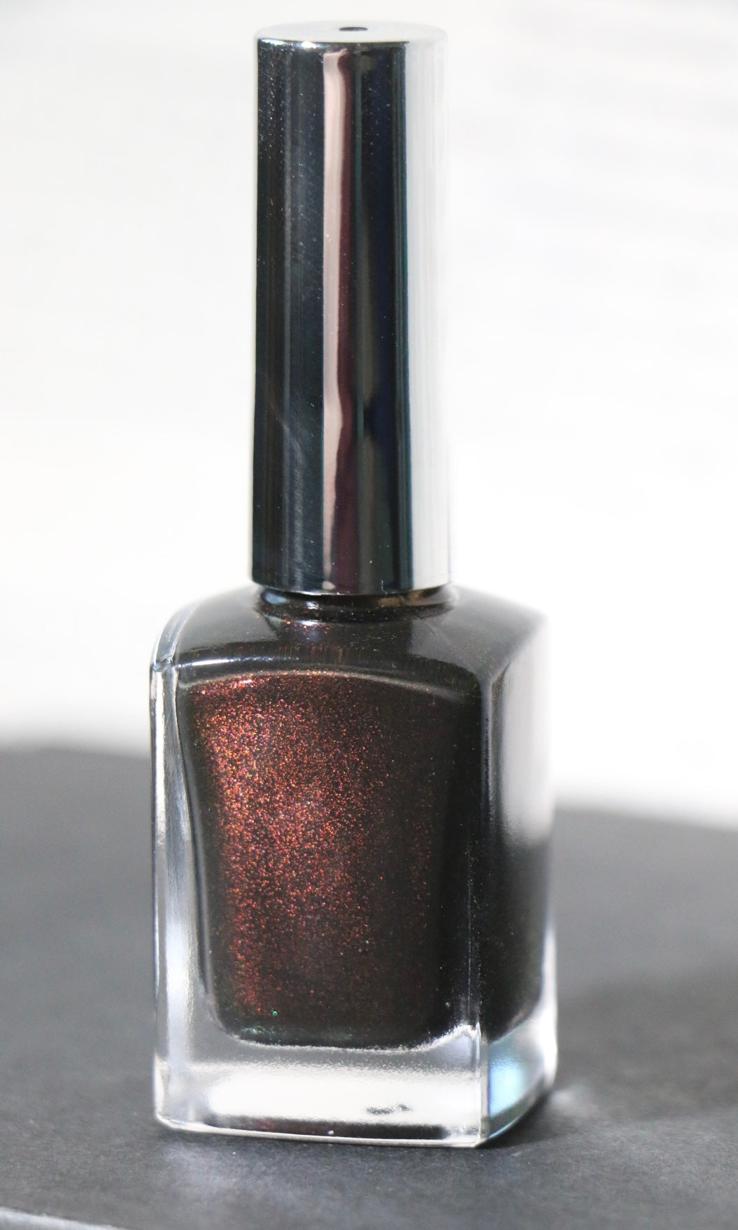 A bottle of dark metallic nail polish with a shimmering bronze-brown finish, featuring a reflective silver cap, set against a light gray background.