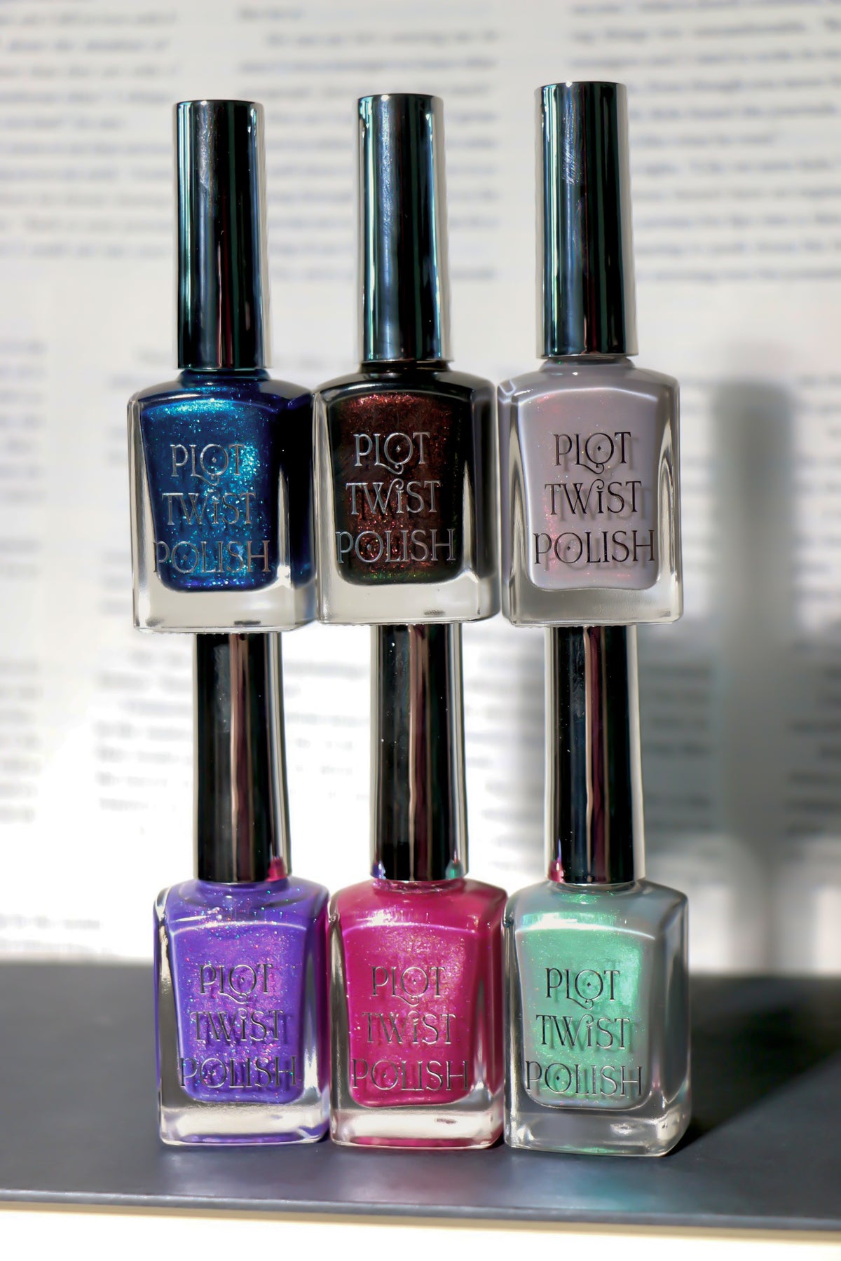 Six bottles of Plot Twist Polish nail polish, showing six different shimmering colors: blue, brown, grey, purple, pink, green. The bottles have silver caps and are set against a blurred background of printed text.