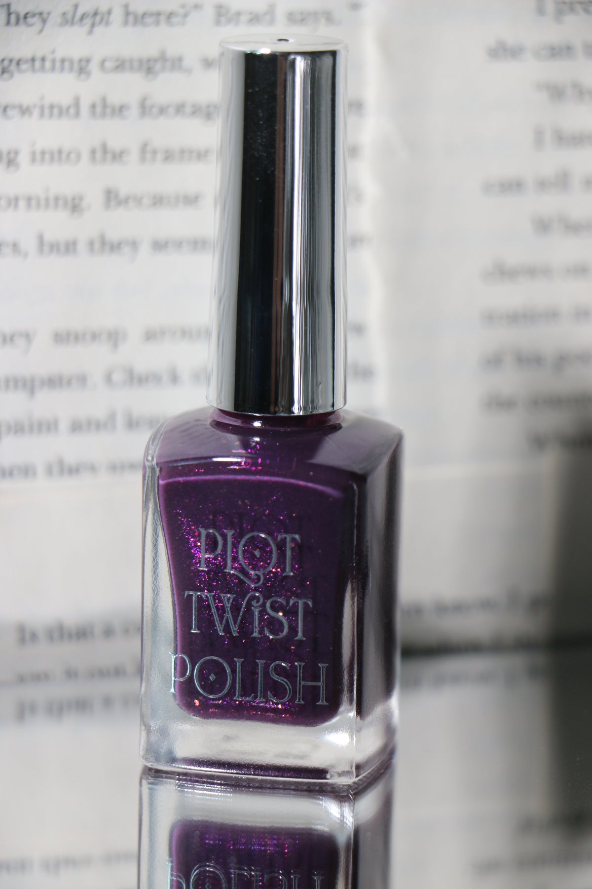 A bottle of Plot Twist Polish nail lacquer in a deep purple shade with silver glitter, featuring a reflective silver cap, set against a blurred background of printed book pages.