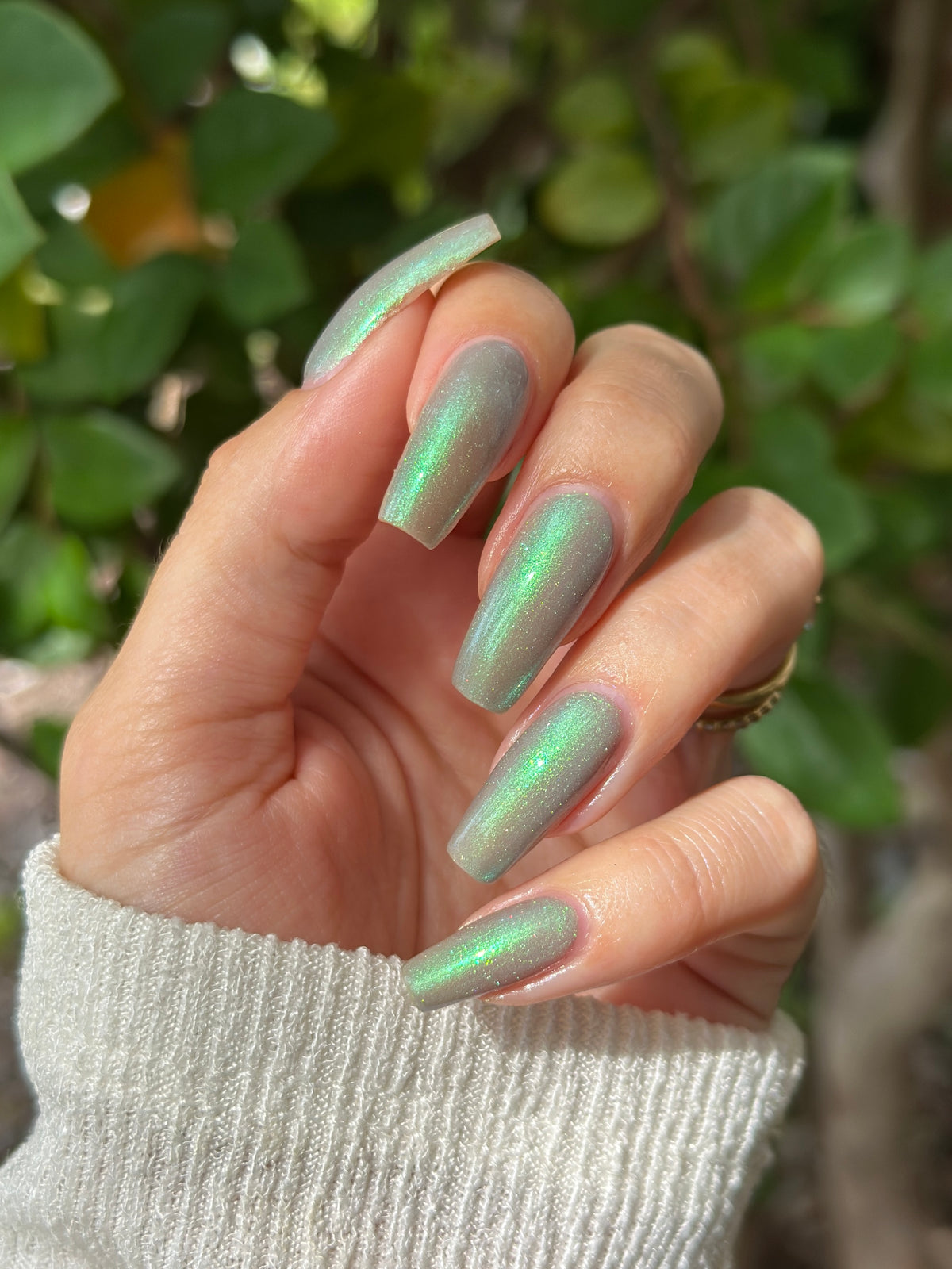 A close-up of a hand with long, almond-shaped nails painted in a green nail polish with a strong iridescent holographic shimmer, set against a blurred background of green foliage.