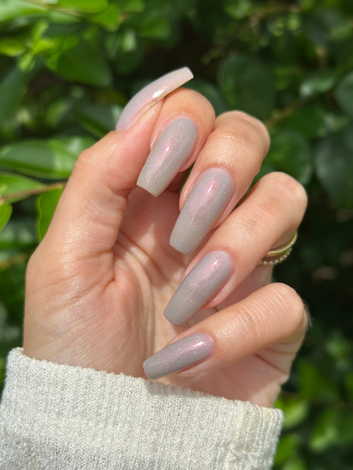 A close-up of a hand with long, almond-shaped nails painted in a sheer, iridescent light gray polish that shifts with a subtle pink shimmer, set against a blurred background of green foliage.