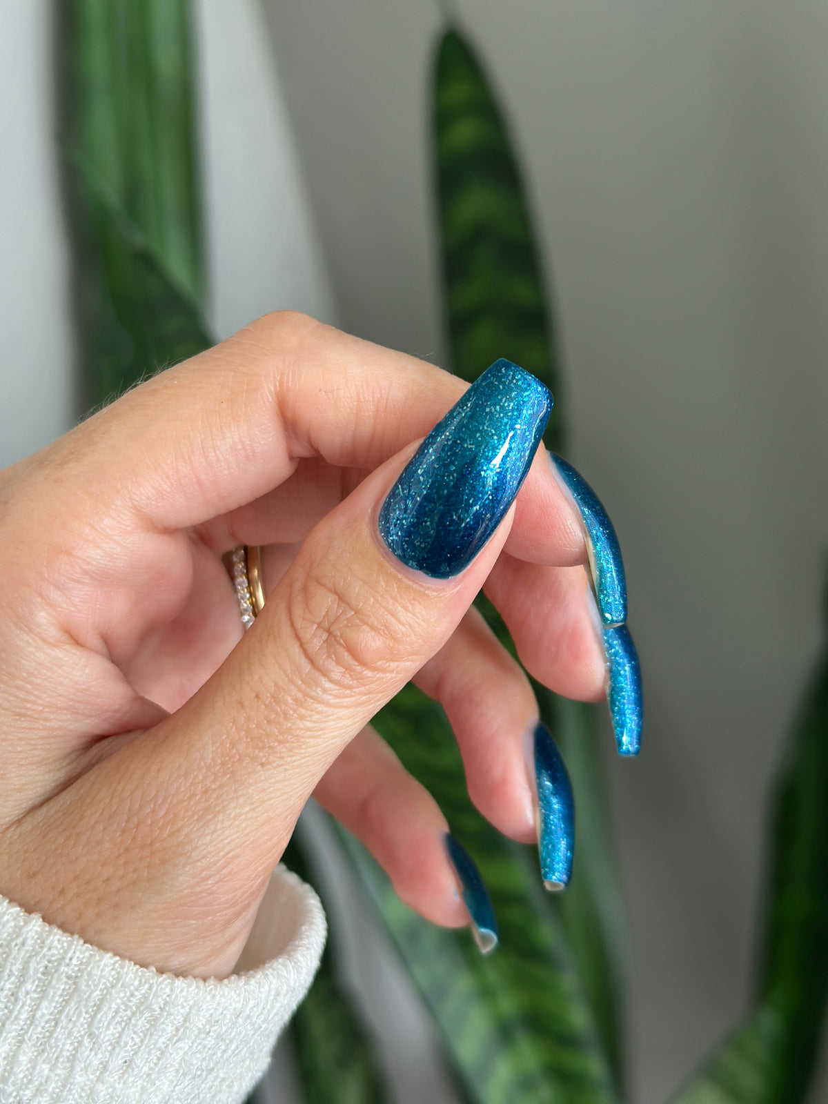 A close-up of a hand with long, sculpted almond-shaped nails painted in a shimmering metallic blue nail polish, with a plant blurred in the background.