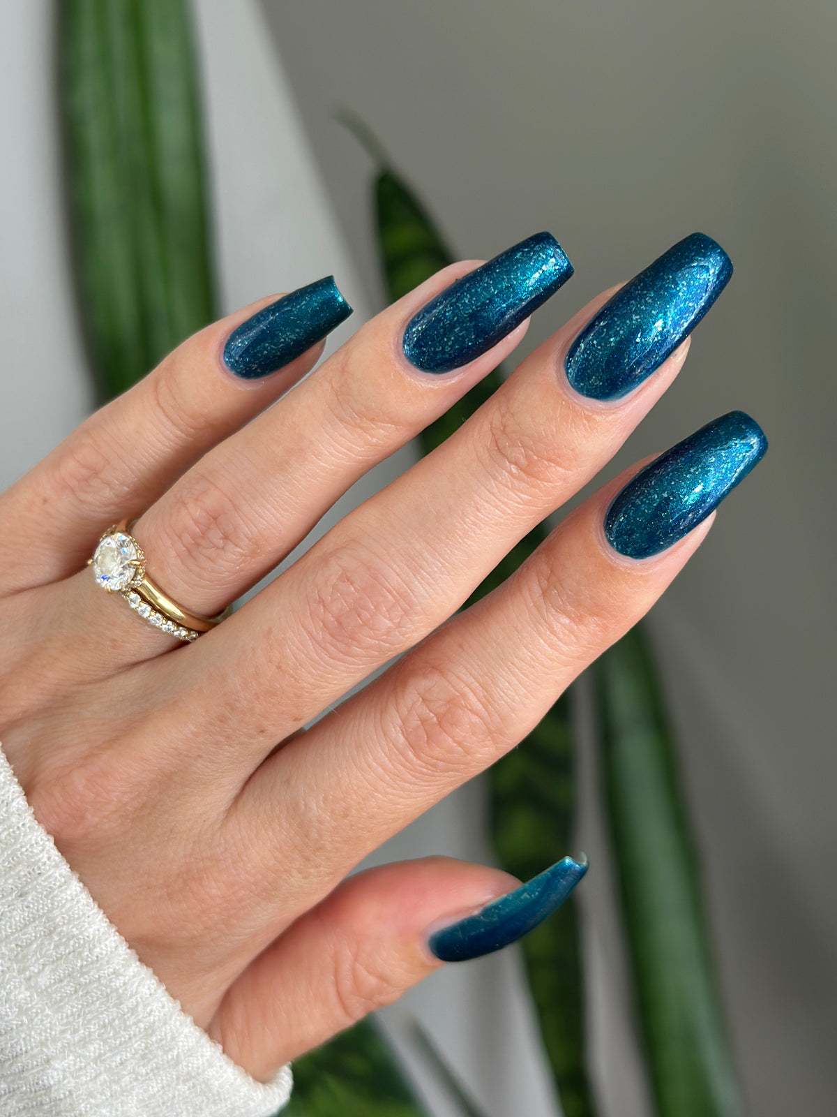 A close-up of a hand with long, coffin-shaped nails painted in a sparkling metallic teal blue nail polish. The hand is wearing a gold and silver ring, and the background features soft, out-of-focus green leaves and a neutral wall.