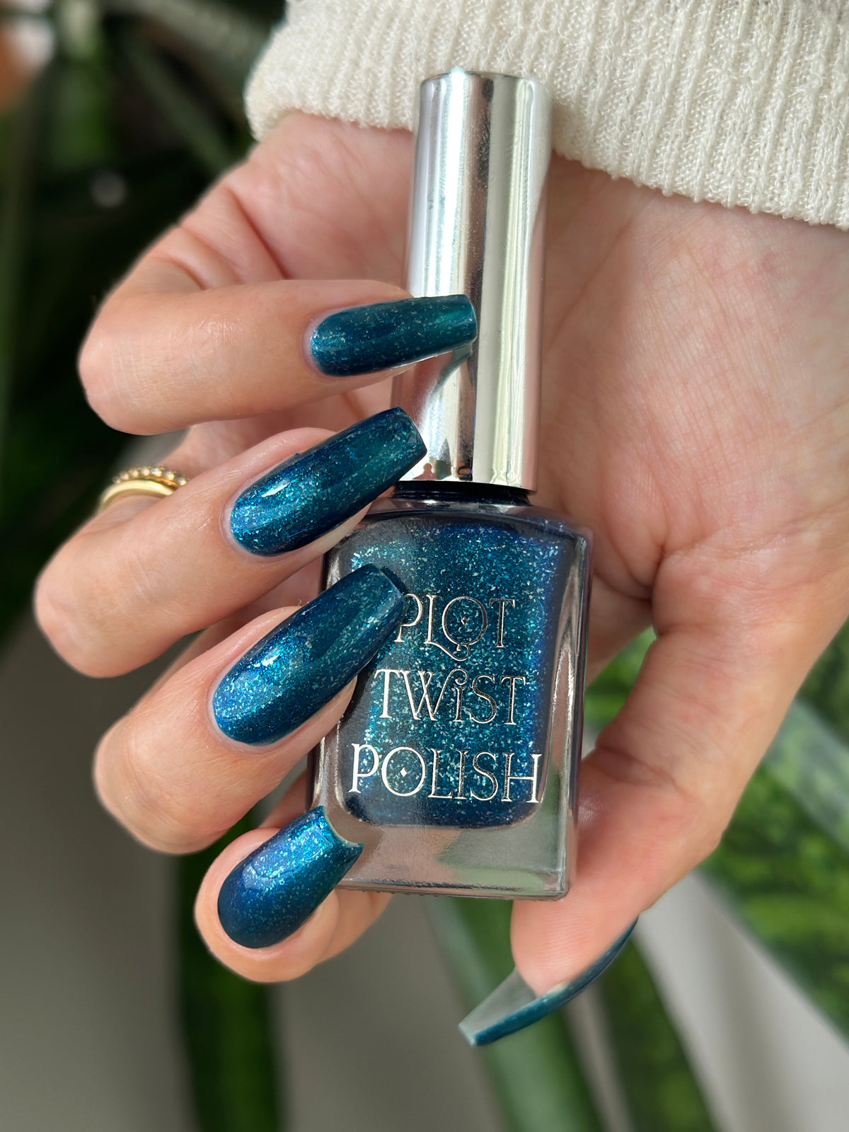 A hand holding a bottle of Plot Twist Polish nail lacquer with a shimmering teal-blue glitter polish, shown against a blurred background.
