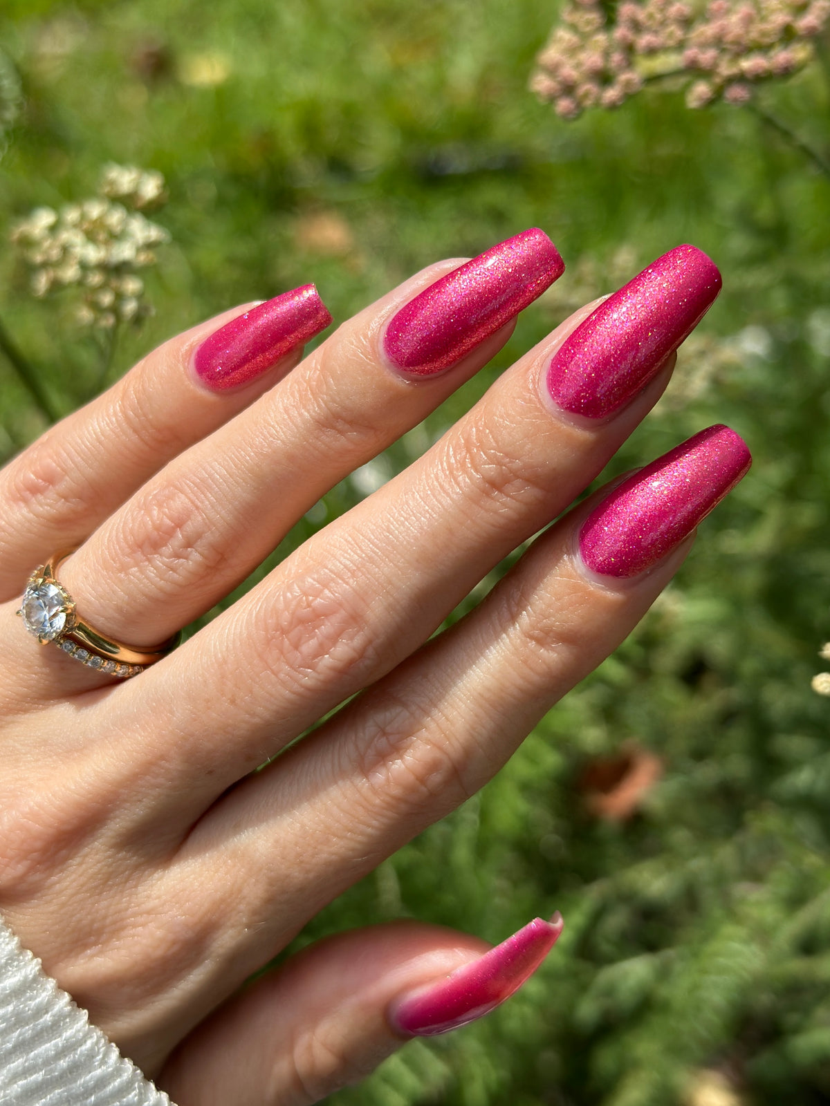 A close-up of a hand with long, glossy, metallic pink manicured nails, wearing a gold ring, set against a blurred green outdoor background.