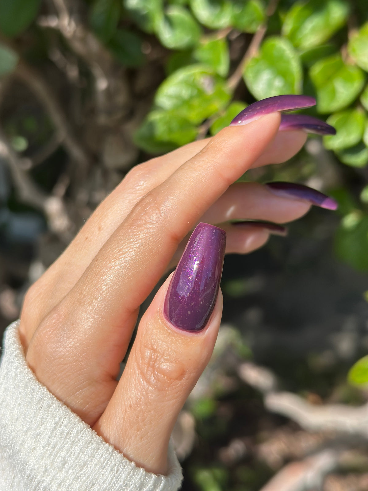 A manicured thumb with a dark purple polish with purple shimmer and small gold flakes