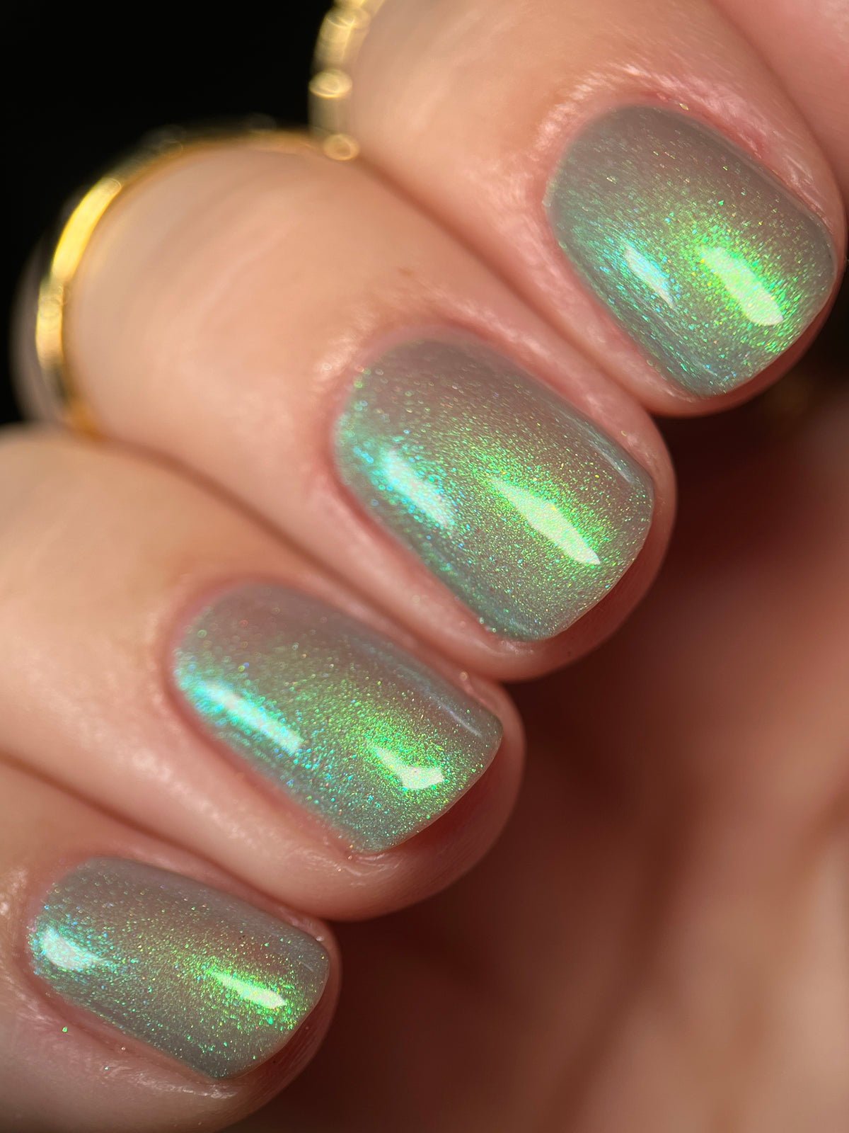 Close-up of a hand with fingernails painted in a shimmering iridescent green nail polish, with a gold ring visible on one finger against a dark background.