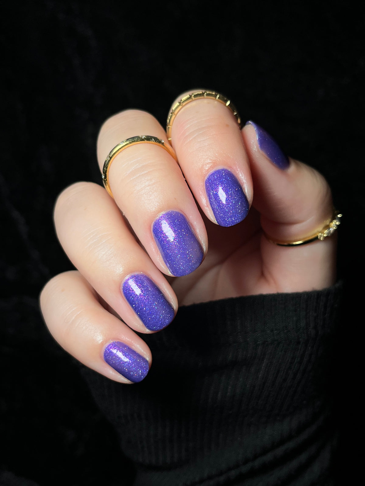 A close-up of a hand with manicured nails painted in a glittery purple shade, wearing gold rings against a black background.