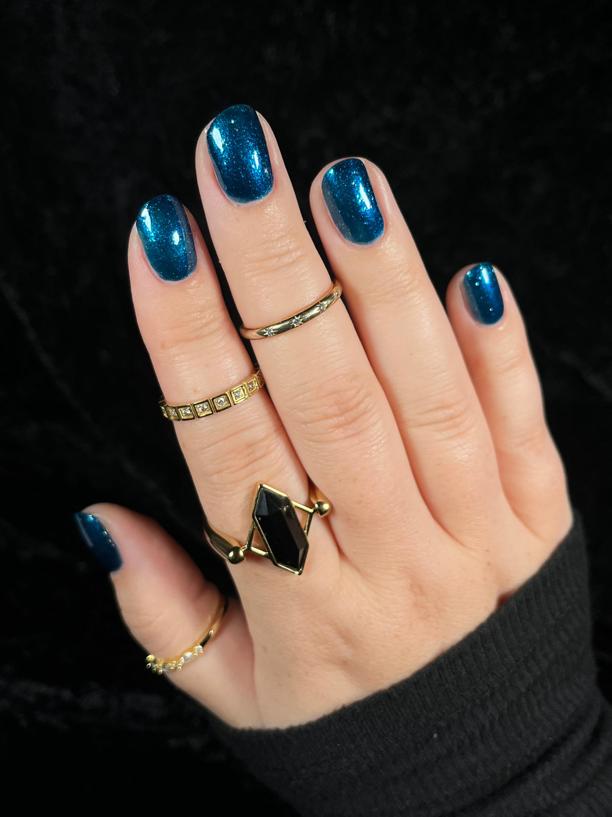 A close-up of a hand wearing multiple gold rings and a blue metallic nail polish against a black background.