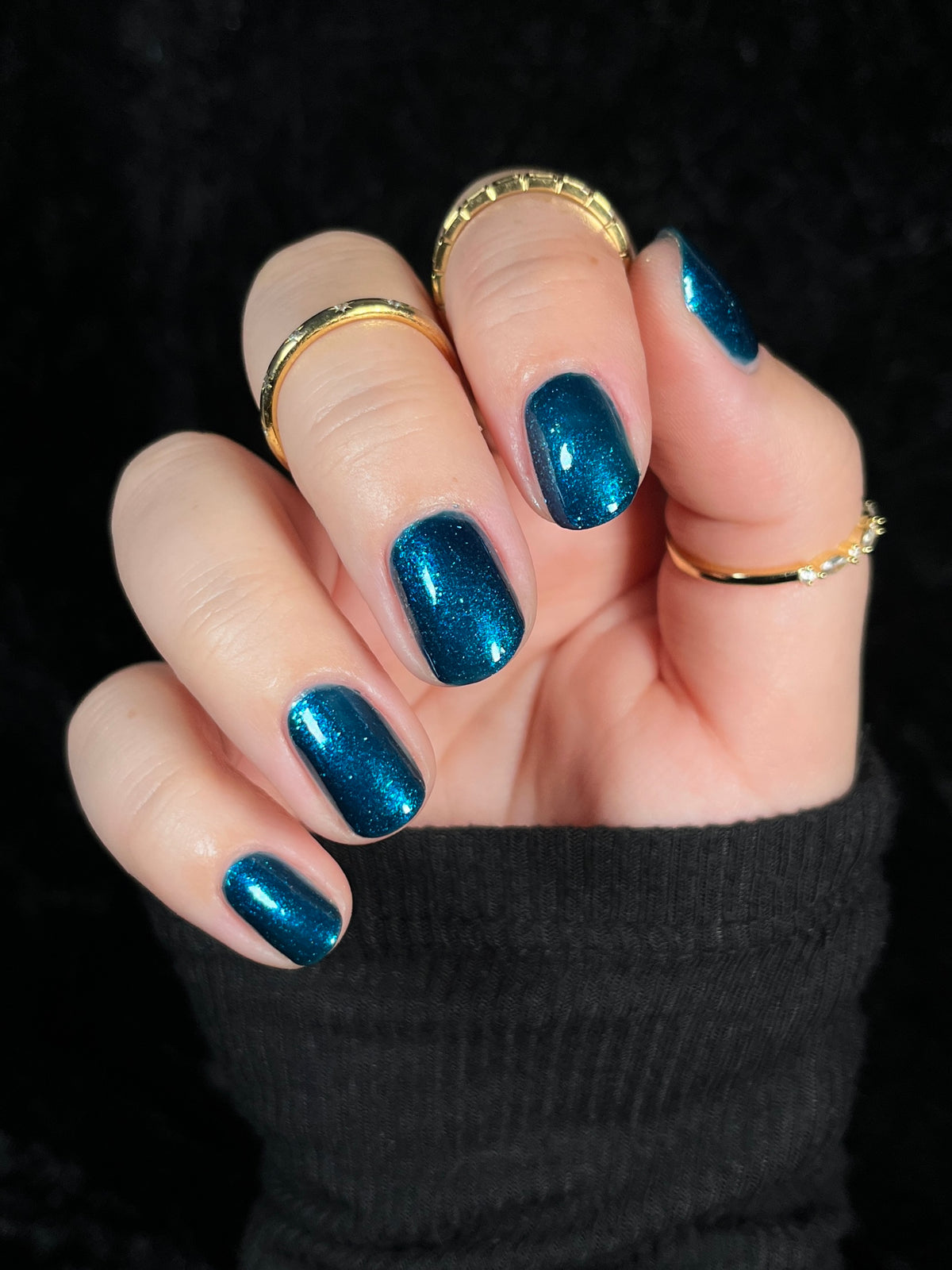 A close-up of a hand with manicured nails painted in a sparkling metallic teal-blue shade, wearing several gold rings against a black background.