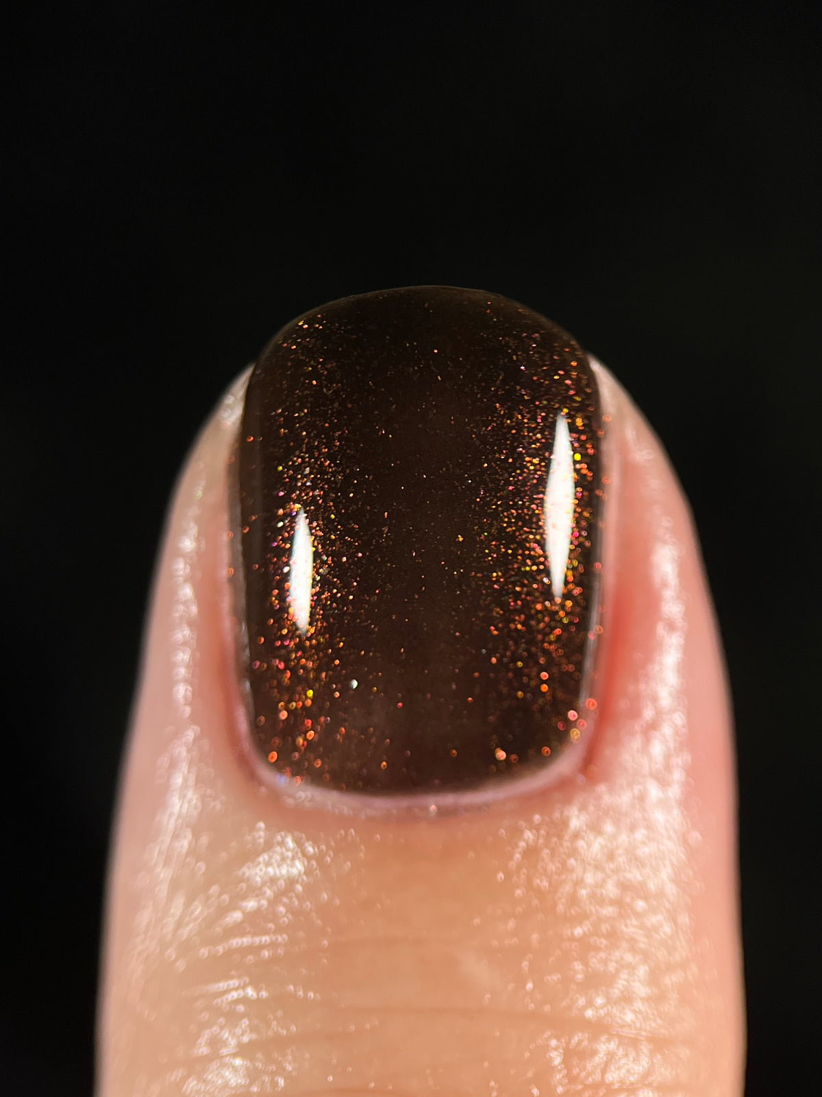 A close-up of a fingernail painted with a glossy dark brown nail polish containing fine shimmering red to orange to gold glitter, set against a solid black background.