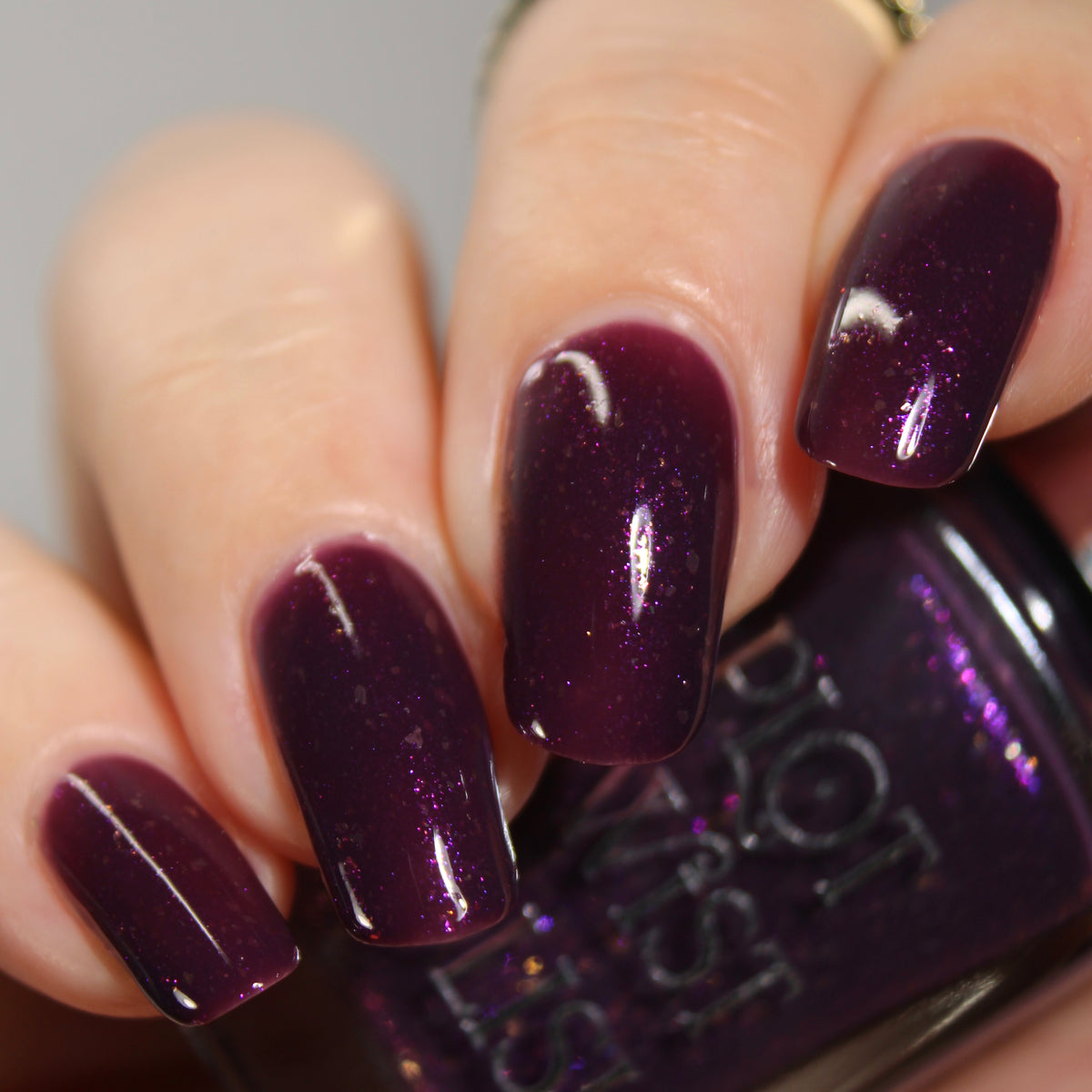 A manicured hand with a dark purple polish with purple shimmer and small gold flakes holding a bottle of nail polish.