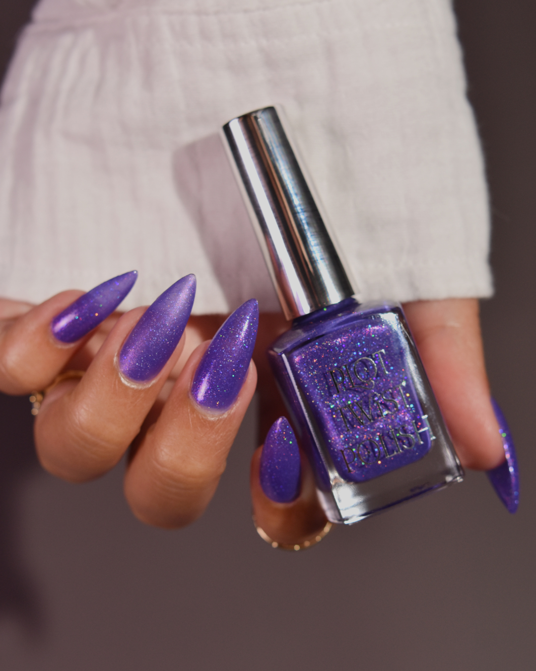 A hand with long, stiletto-shaped nails painted in a sparkling purple polish, holding a bottle of purple glitter nail polish against a soft neutral background.