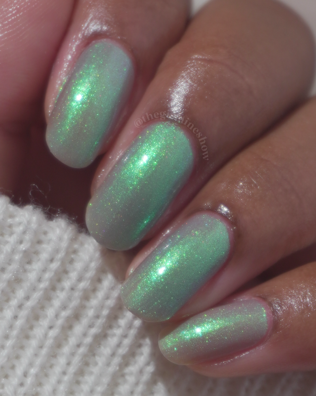 Close-up of a hand with manicured nails painted in a shimmering green and iridescent holographic nail polish, resting on a white knit fabric.