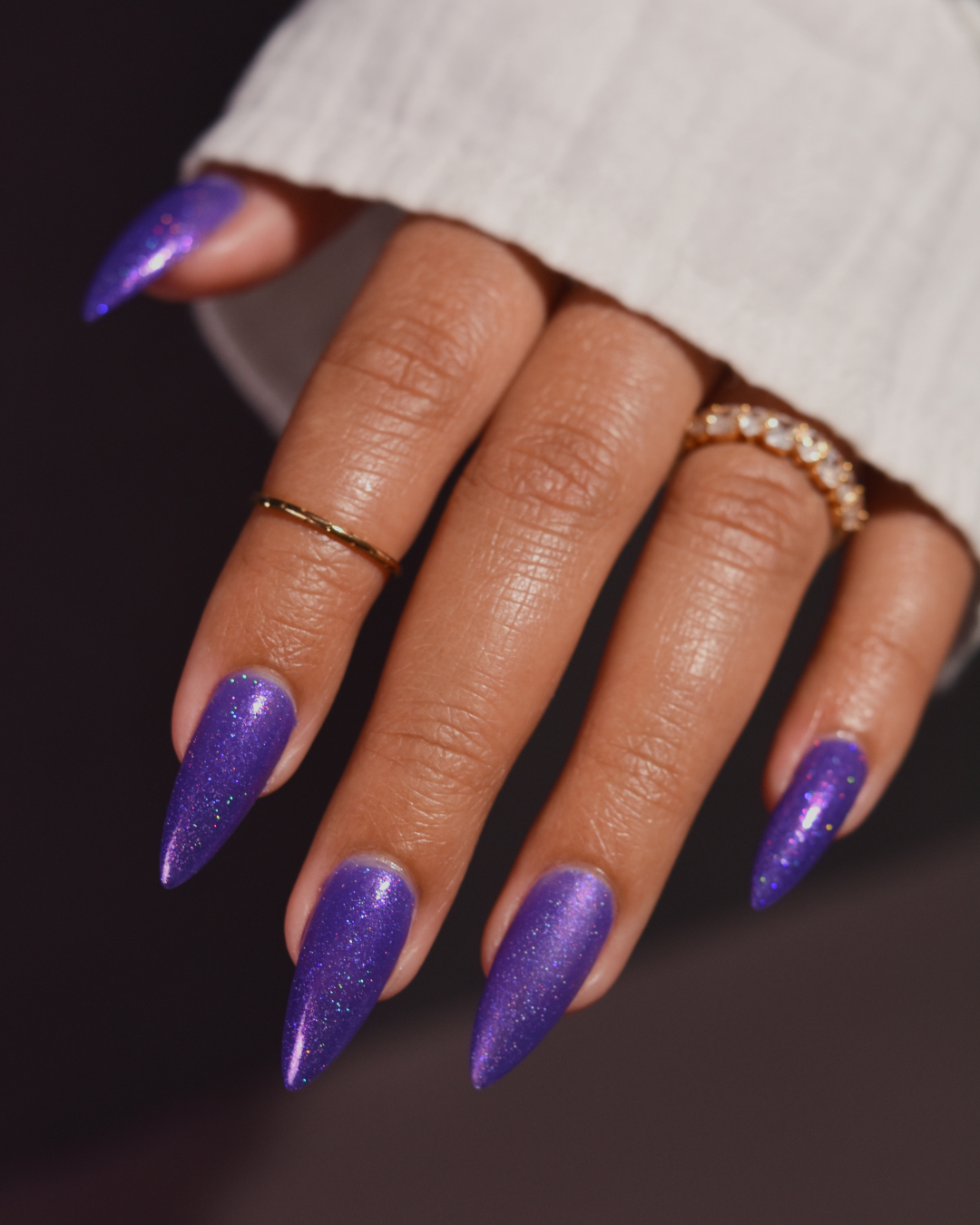 A close-up of a hand with long, stiletto-shaped nails painted in a sparkling purple glitter polish, wearing a white ribbed sweater and gold rings against a dark background.