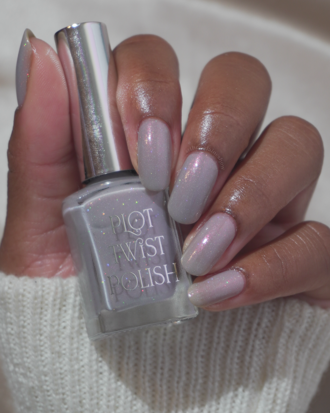 A close-up of a hand with long, glossy, grey manicured nails holding a bottle of Plot Twist Polish.