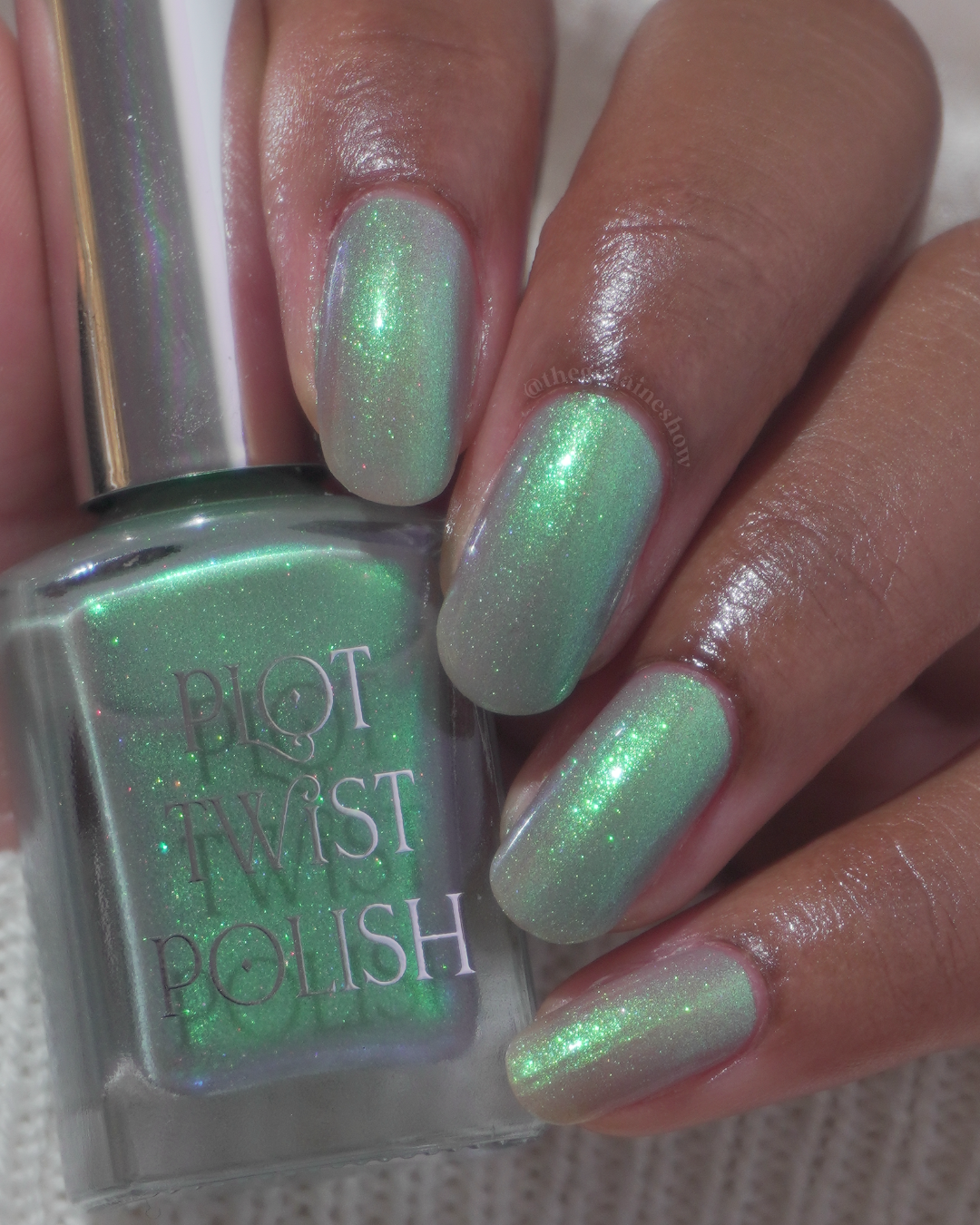 A close-up of a hand with nails painted in a shimmering green nail polish, holding a bottle of Plot Twist Polish in the same shade.