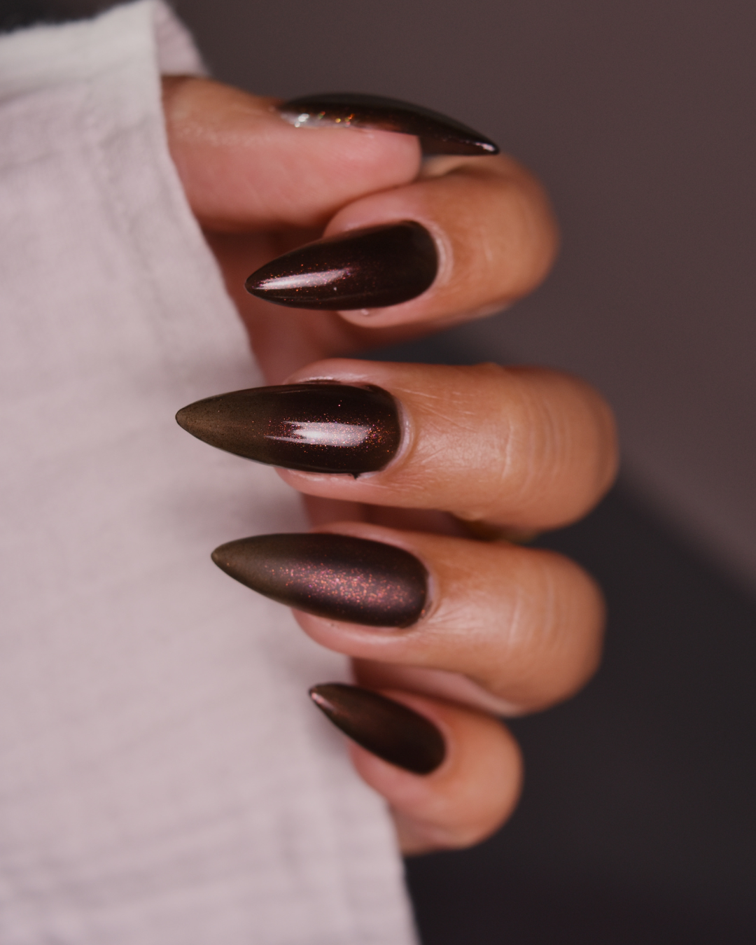 A close-up of a hand with long, stiletto-shaped nails painted in a deep metallic espresso brown with subtle shimmer, holding a white textured fabric against a dark gray background.