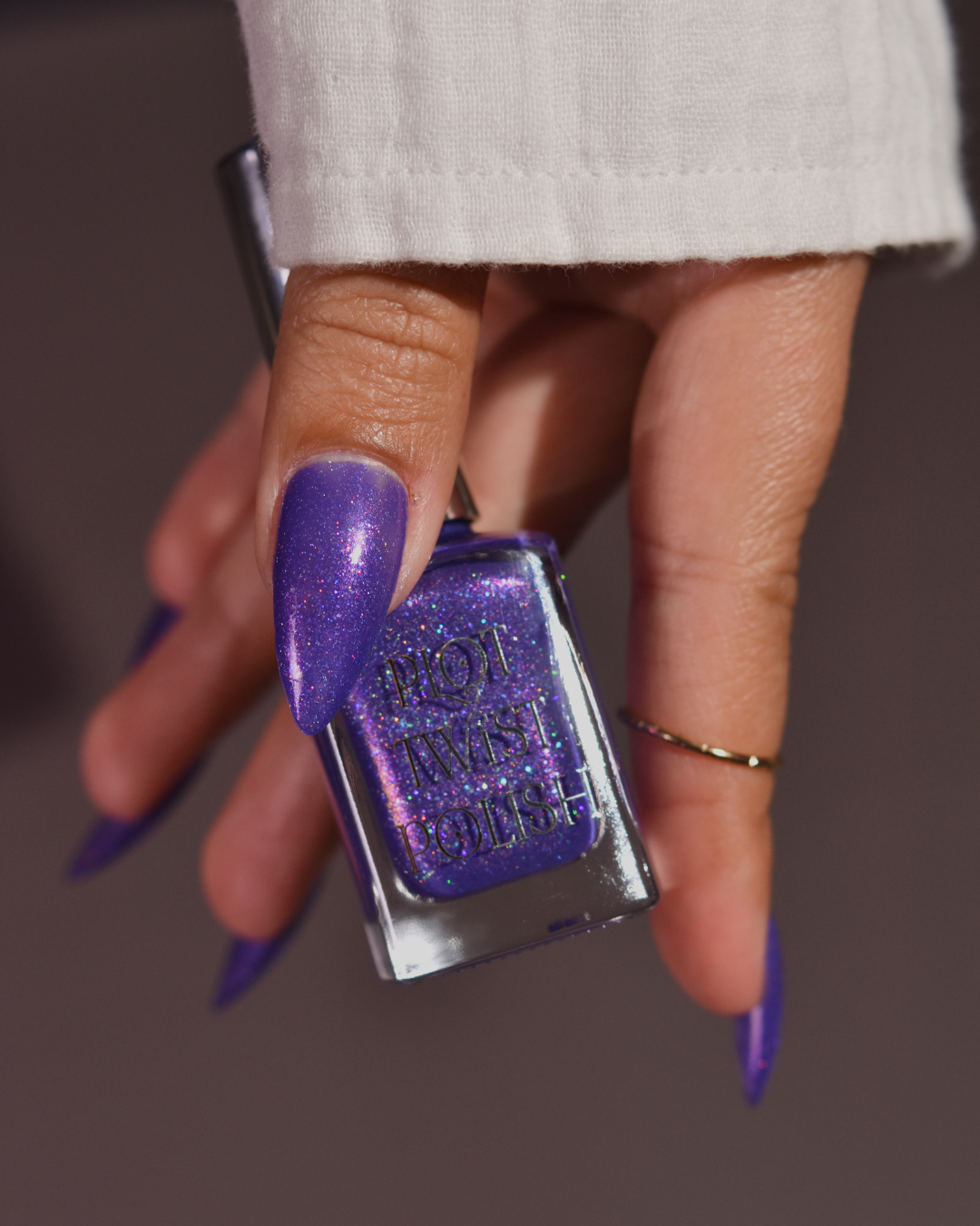 A hand with long, manicured nails painted in a shimmering purple nail polish, holding a bottle of 'Pilot Twist Purple' nail polish.