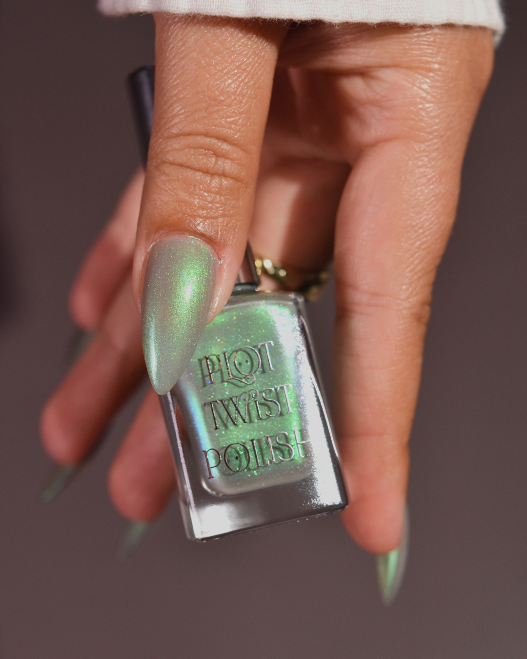 A hand holding a bottle of Plot Twist Polish nail polish in a shimmering light green shade, with the manicured nails painted in the same color against a dark gray background.