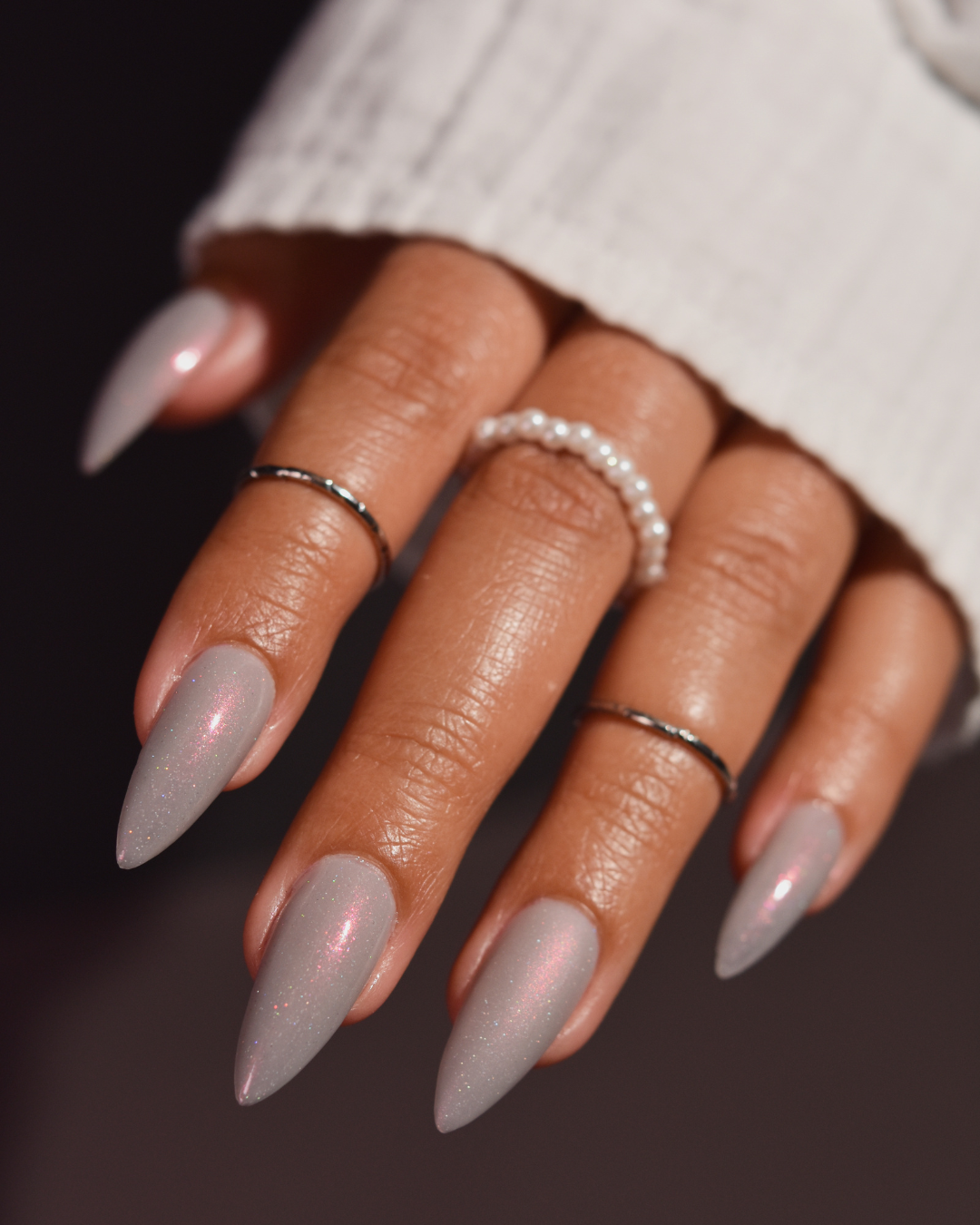 A close-up of a hand with long, stiletto-shaped nails painted in a sheer, glittery grey shade. 