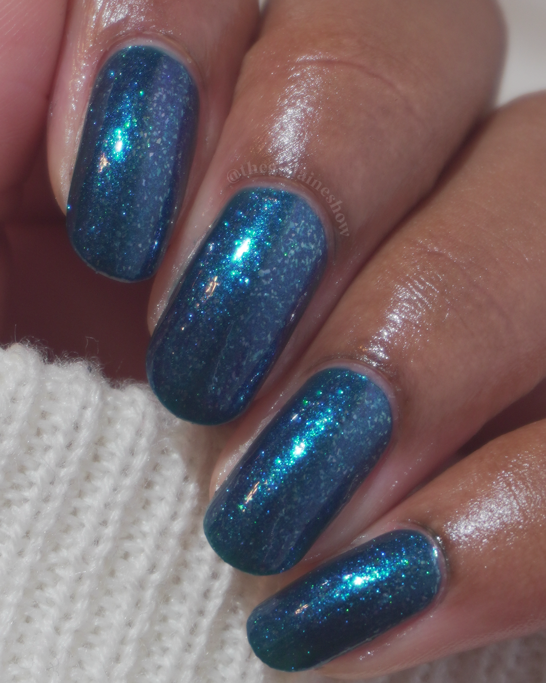 Close-up of a hand with fingernails painted in a deep teal-blue glitter polish, resting against a white knit fabric.