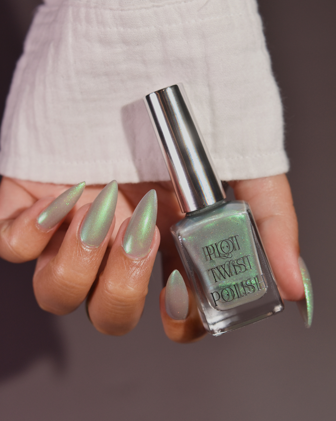 A hand with long stiletto nails painted in a shimmering green nail polish, holding a bottle of Plot Twist Polish against a dark gray background.
