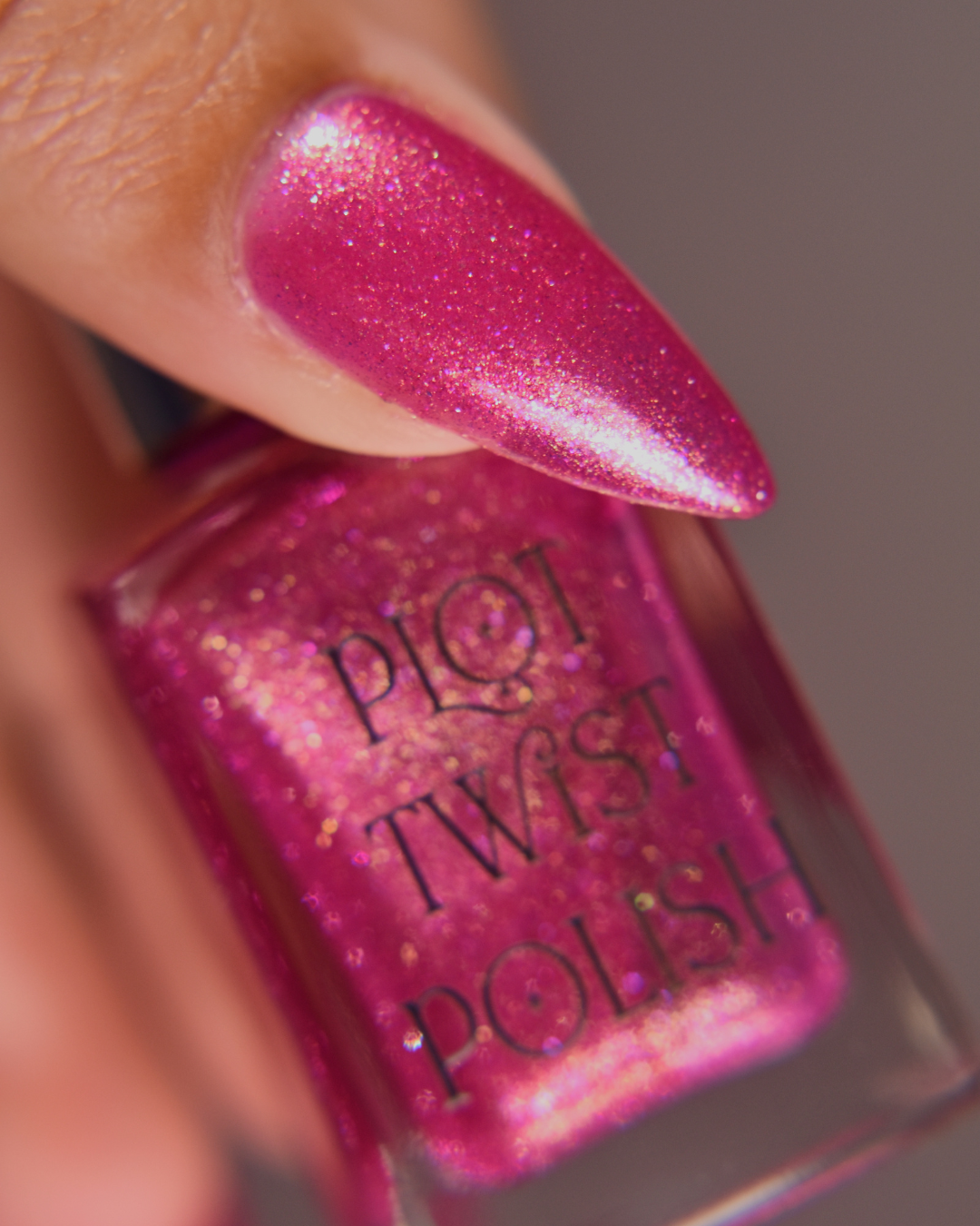 A close-up of a hand with long stiletto nails painted in a sparkling magenta pink nail polish, holding a bottle of Plot Twist Polish in the same shade.