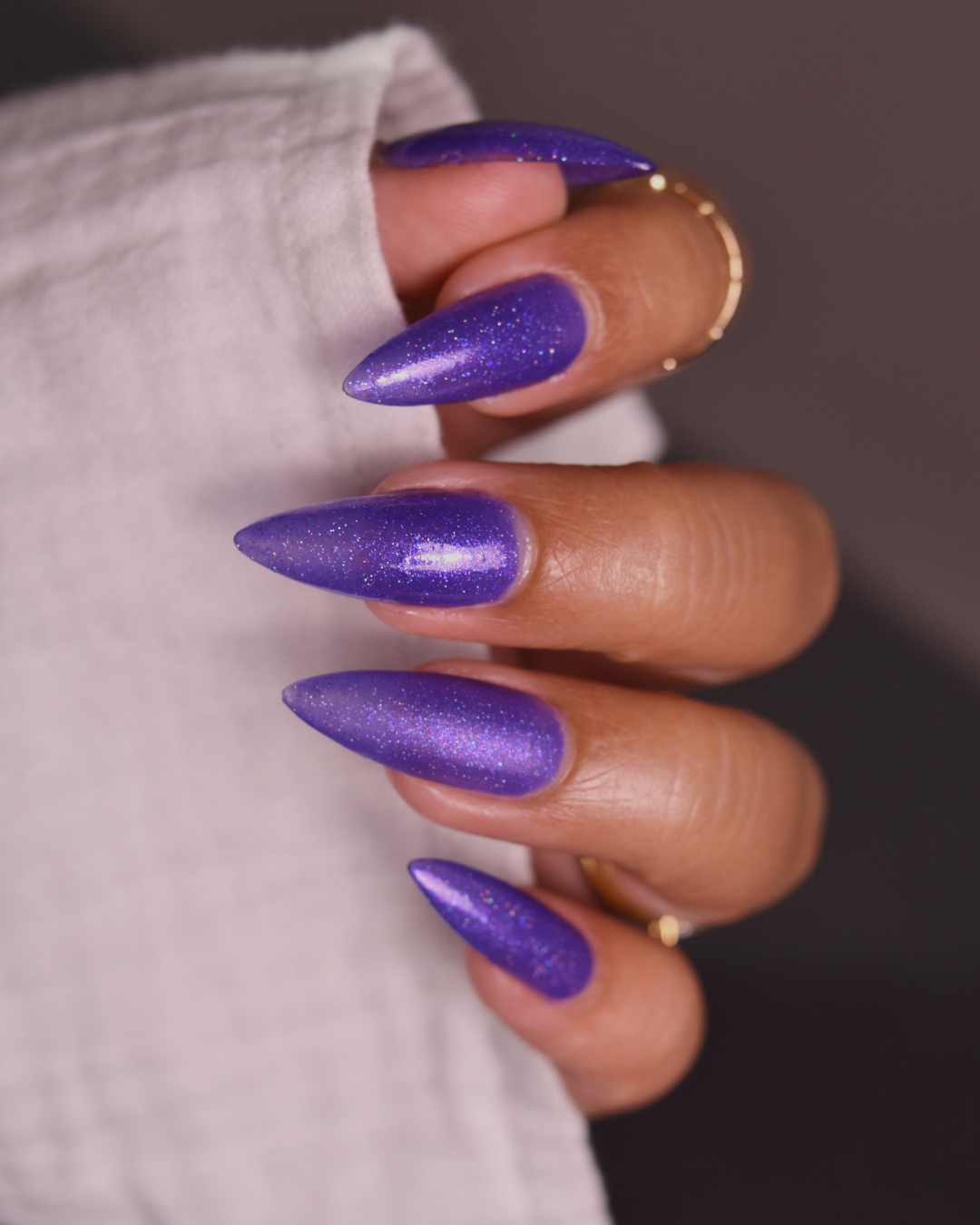 A close-up of a hand with long, stiletto-shaped nails painted in a sparkling metallic purple shade, resting against a light fabric background.