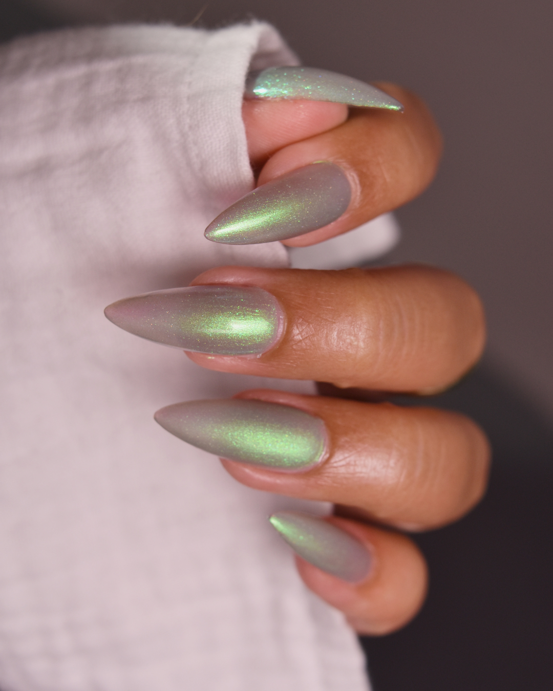 A close-up of a hand with long, stiletto-shaped nails painted in a shimmering iridescent green color, resting against a white textured fabric.