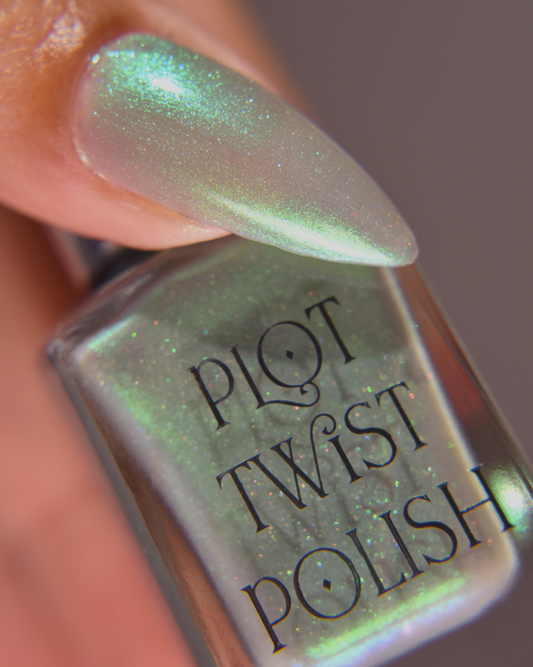 A close-up of a fingernail painted with iridescent mint green nail polish, holding a bottle labeled 'PLOT TWIST POLISH'.