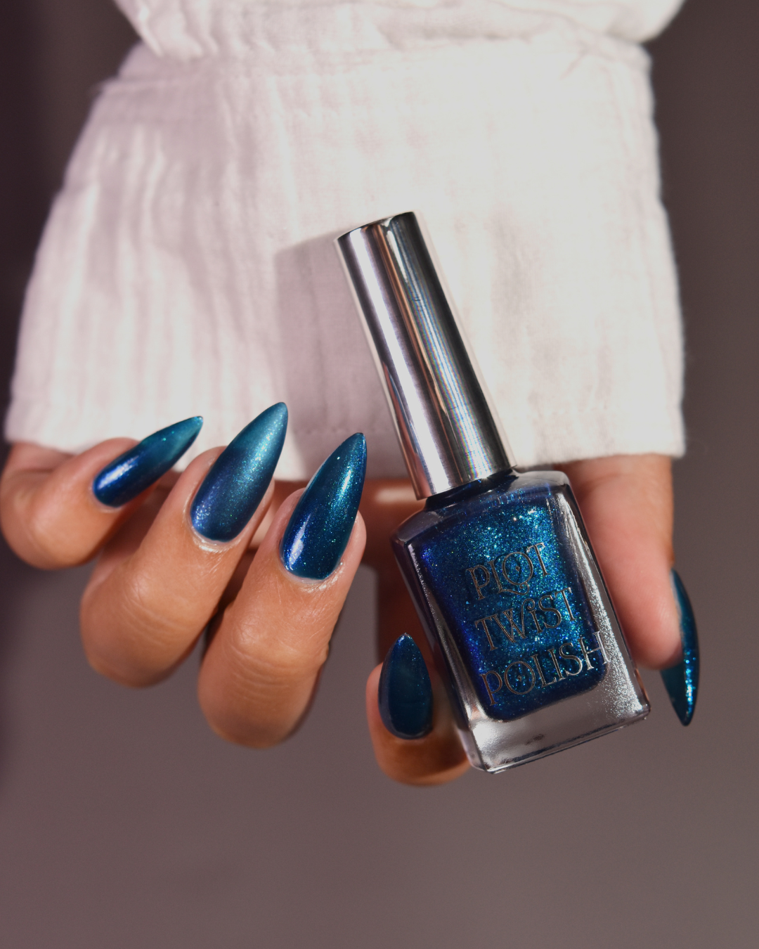 A hand with long, metallic teal-blue manicured nails holds a bottle of Plot Twist Polish nail polish. The bottle is clear glass with a silver cap and shows the blue glitter polish inside.