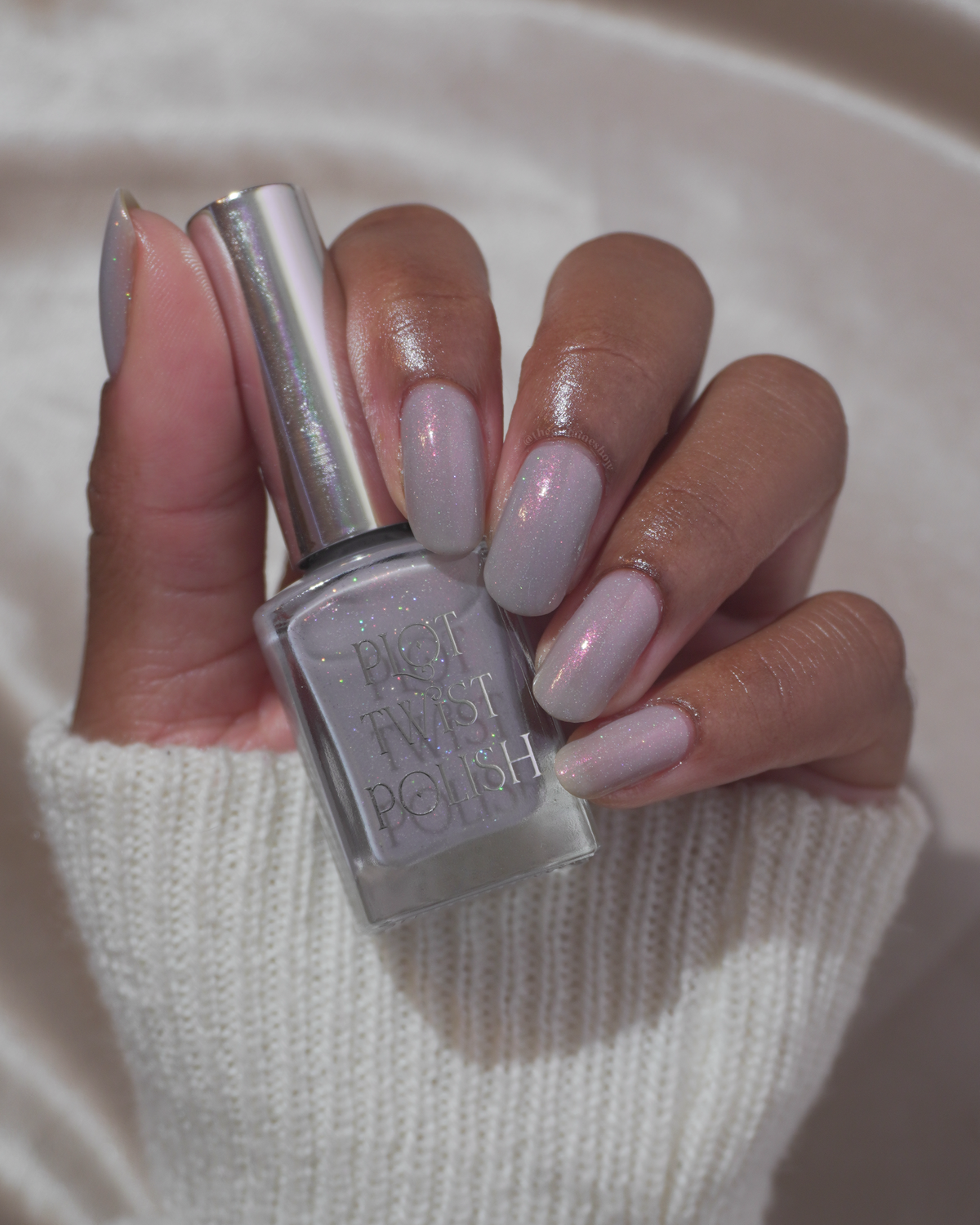A hand holding a bottle of nail polish with matching light grey glitter nail polish applied, against a soft cream-colored background.