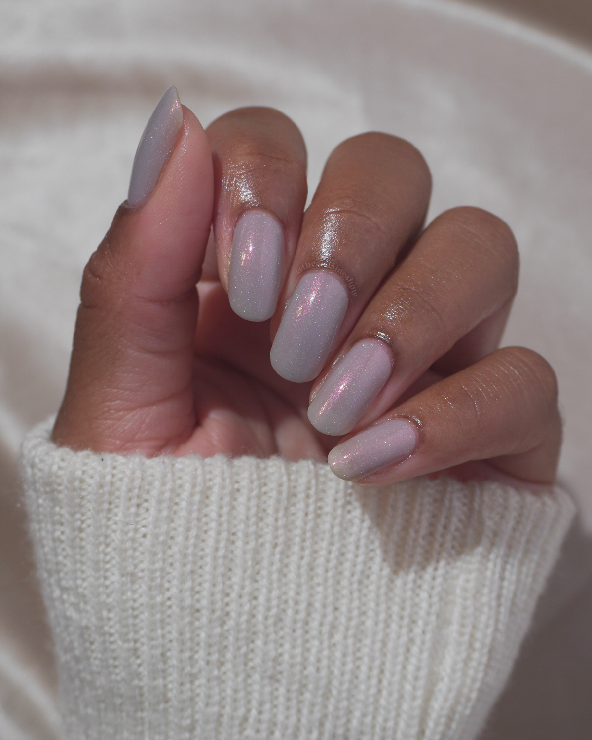 A close-up of a hand with manicured nails painted in a glossy gray color with a subtle shimmer, wearing a white ribbed knit sweater.
