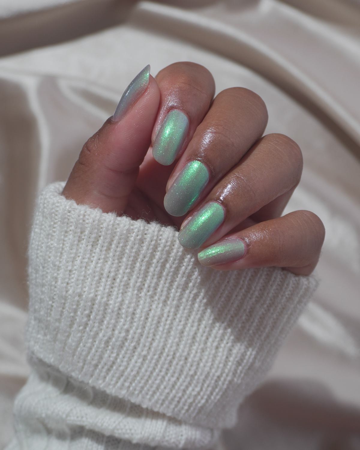 A close-up of a hand with manicured nails painted in a glossy green shade, wearing a white ribbed knit sweater cuff against a soft beige fabric background.