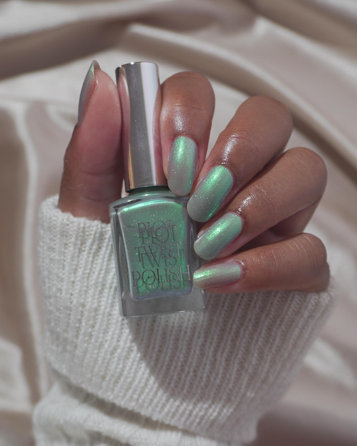 A hand holding a bottle of Plot Twist Polish nail lacquer in a shimmering green shade, with the nails painted to match, set against a soft beige knit sweater and draped fabric background.