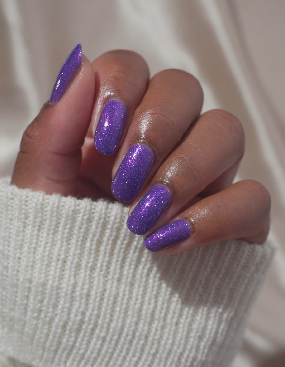 A close-up of a hand with long, almond-shaped nails painted in a sparkling purple glitter nail polish, resting on a white ribbed knit sleeve against a soft white fabric background.