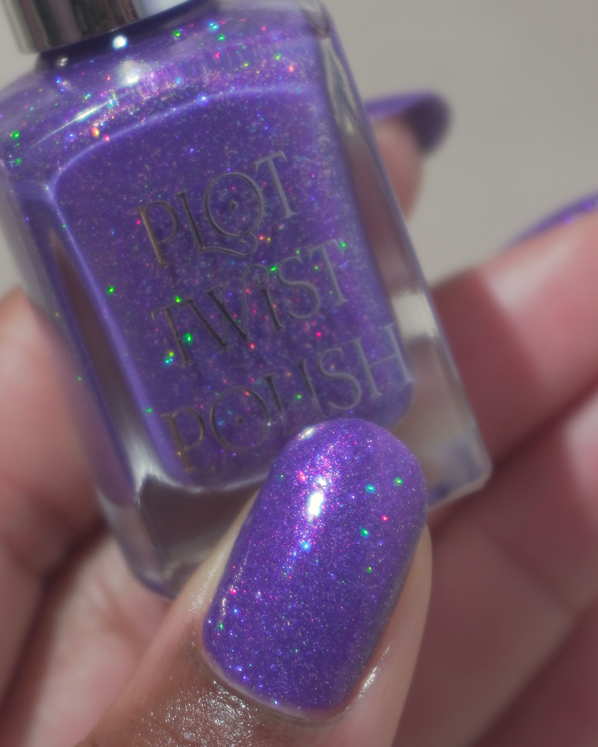 A close-up of a hand holding a bottle of Plot Twist Polish in a sparkling purple shade, with the matching purple nail polish applied to the fingernail.